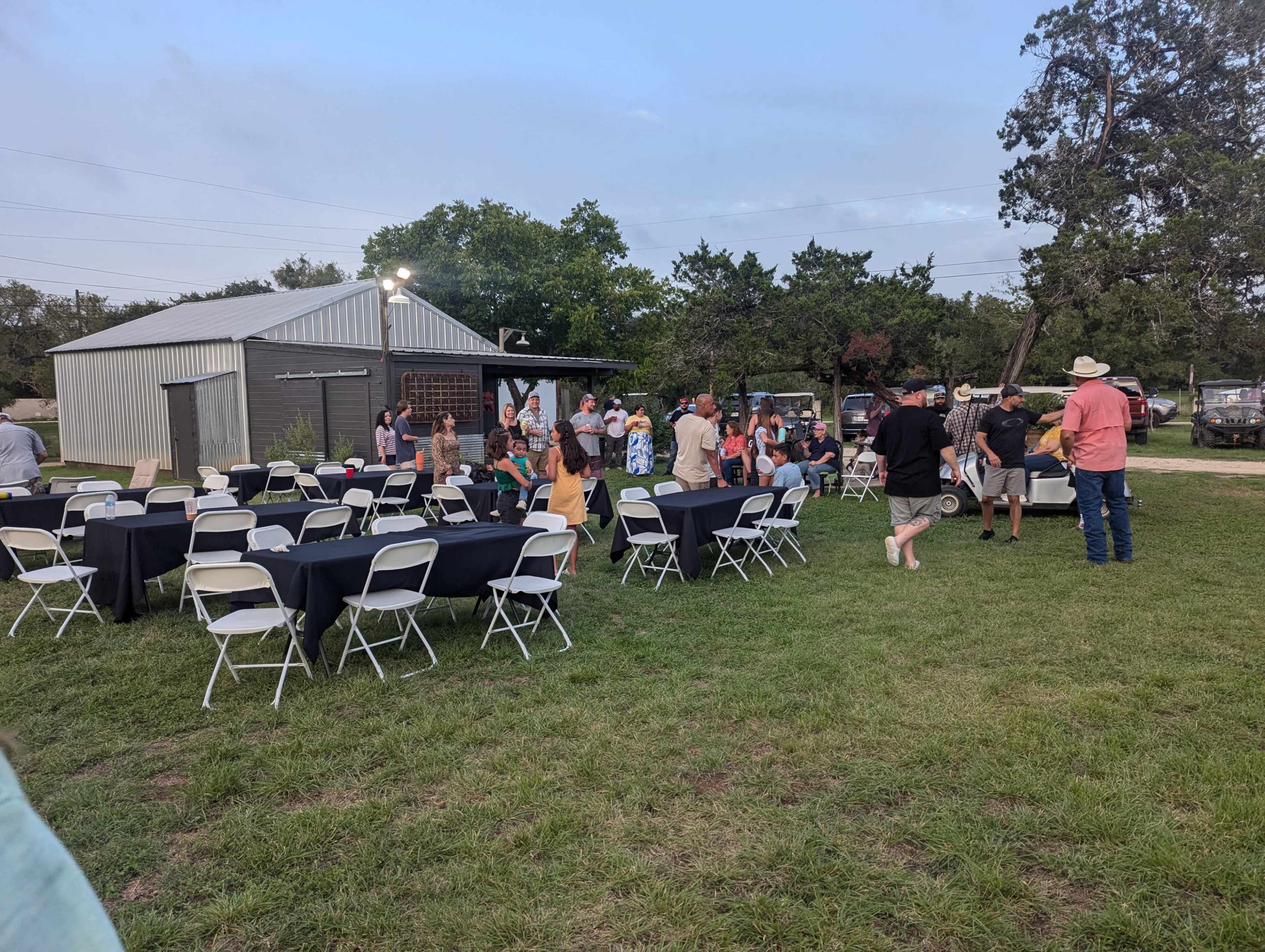 A gathering of people is taking place outdoors near a barn, with tables set up around a grassy area.