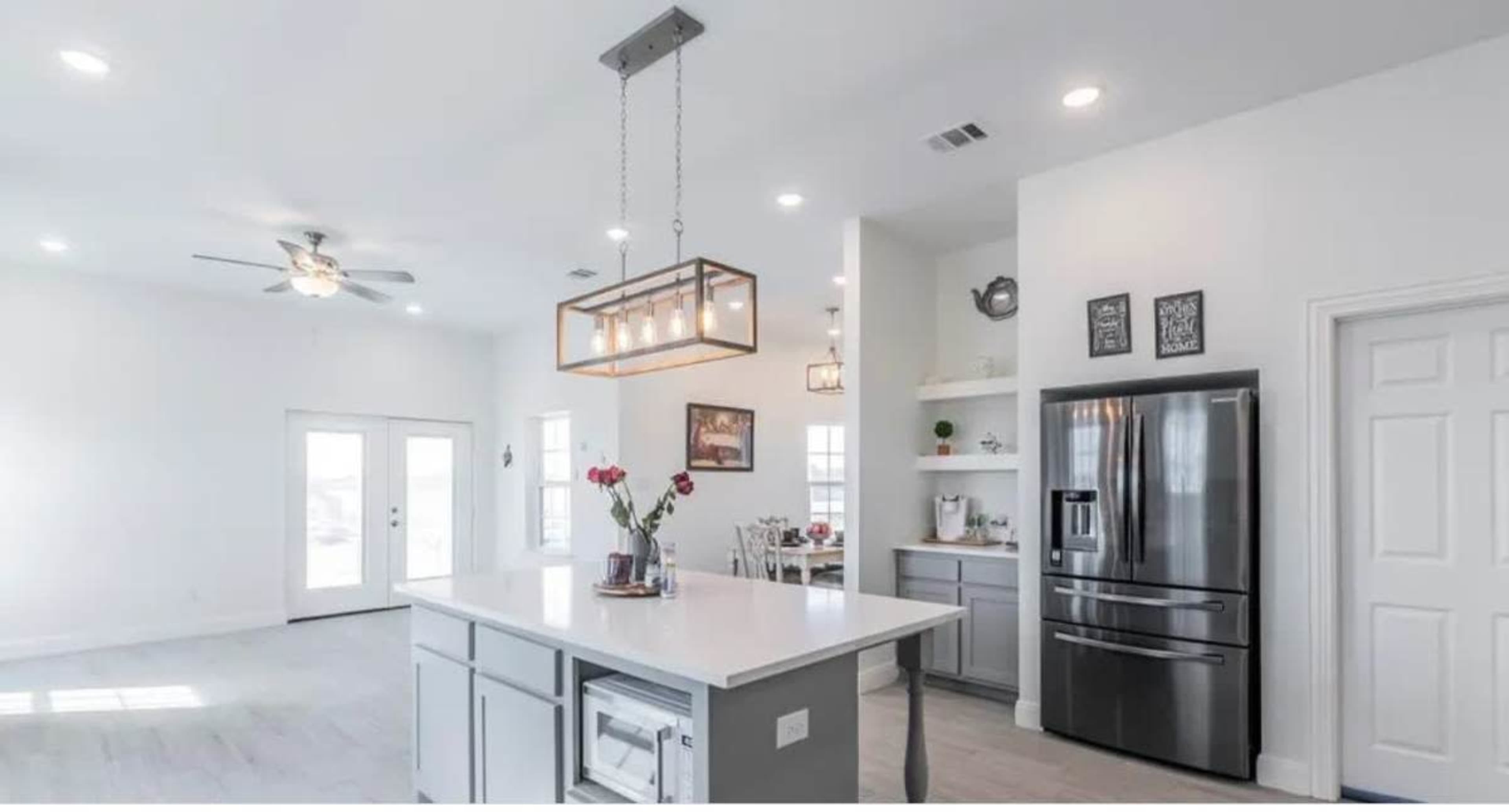 The image shows a modern kitchen with gray cabinetry, a large island with a white countertop, and a stainless steel refrigerator, featuring bright lighting and an open layout.