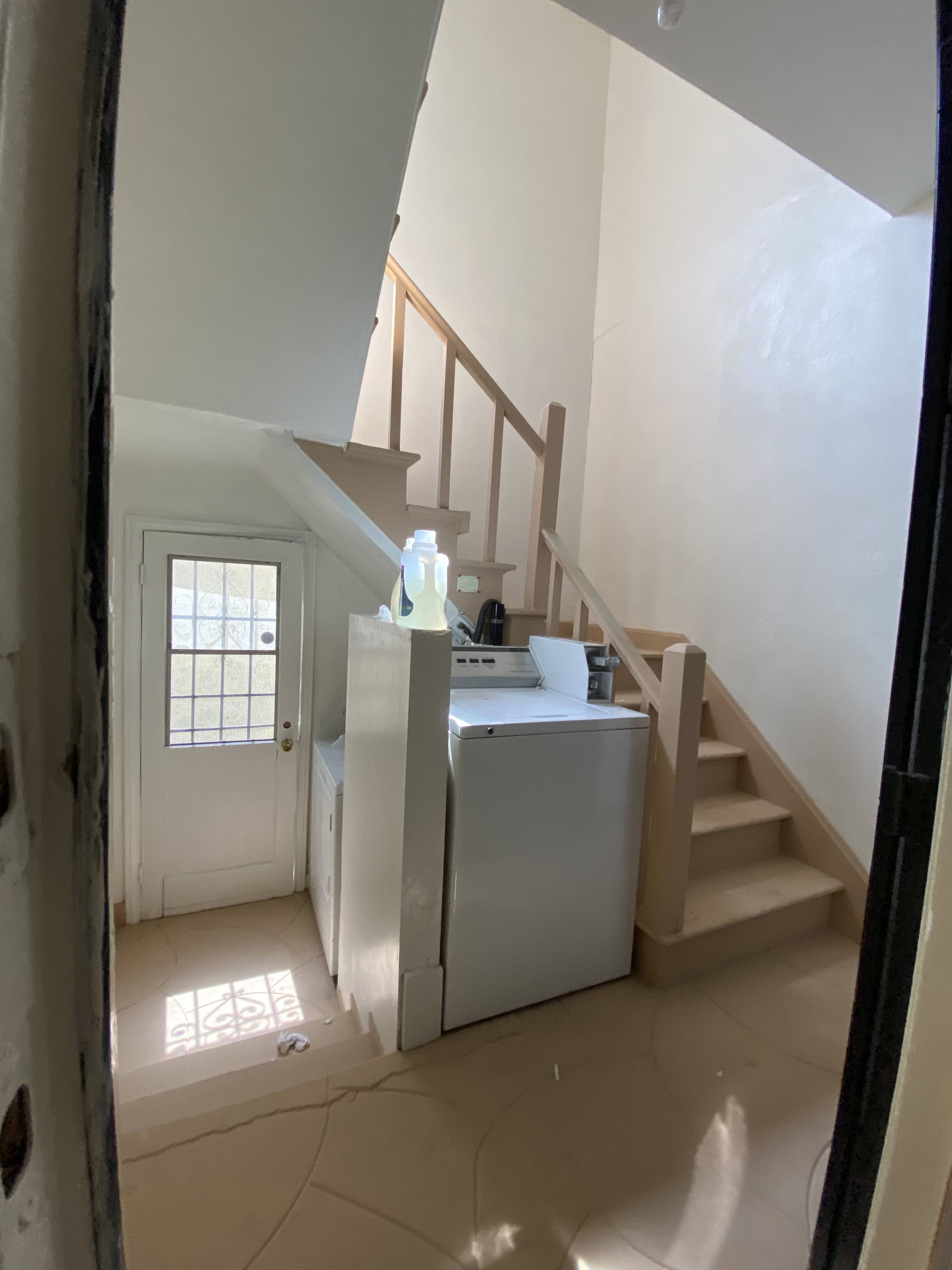The image shows a stairwell with a washing machine and dryer situated near the bottom, adjacent to a door leading outside.