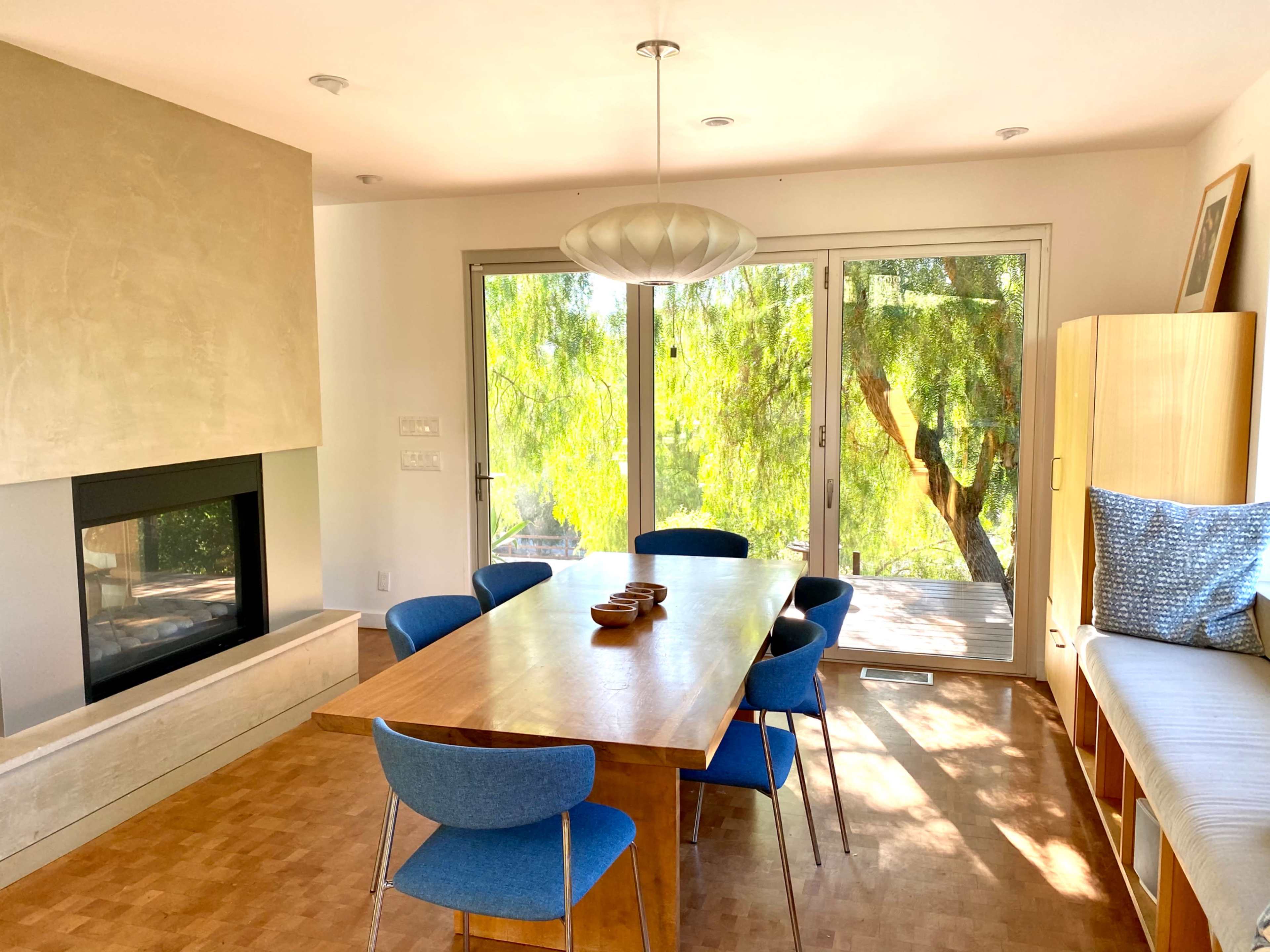 A dining room features a wooden table surrounded by blue chairs, with large windows showcasing natural greenery outside.