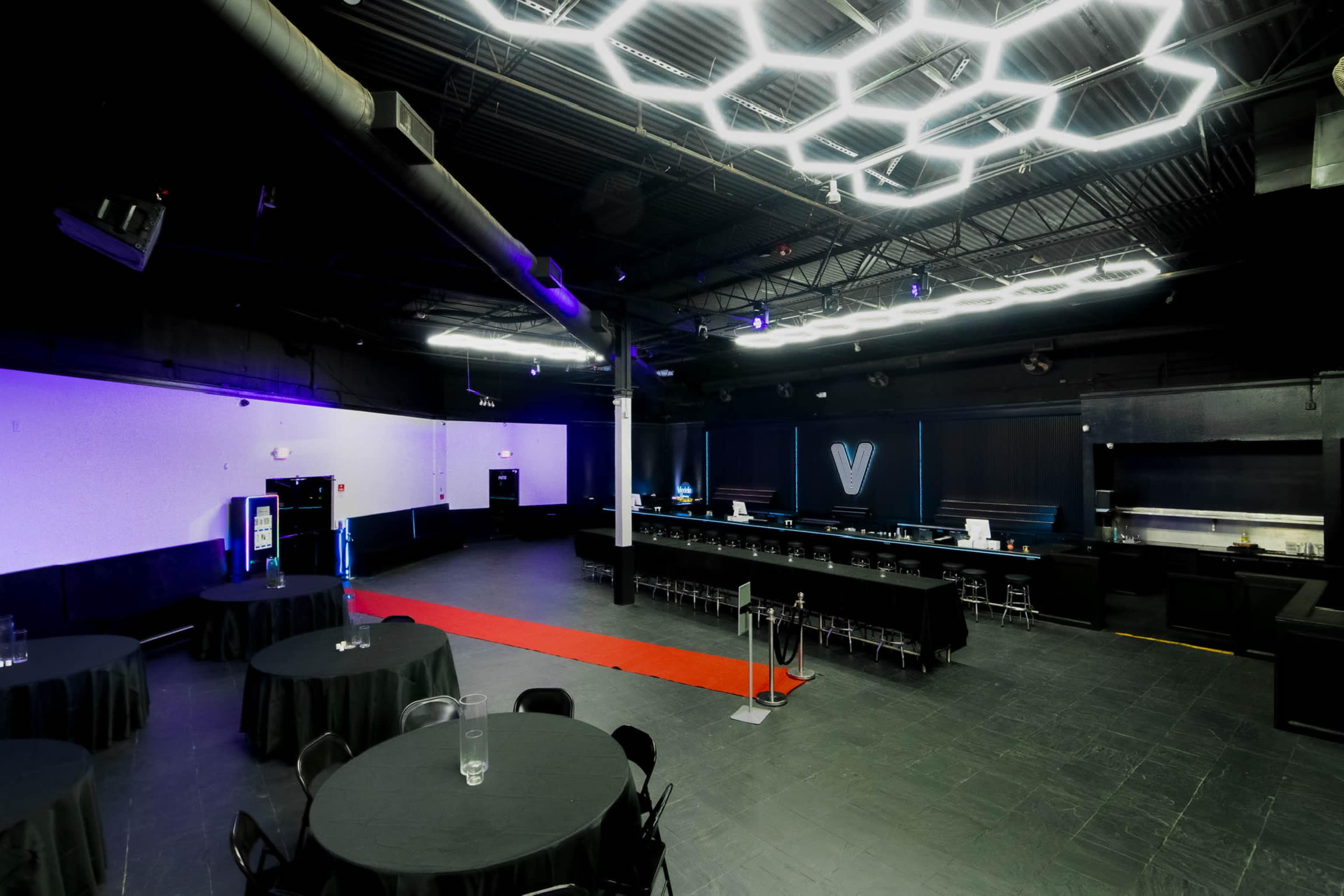 The image shows a large, dimly lit event space with black tables, a red carpet, and a hexagonal light fixture on the ceiling.