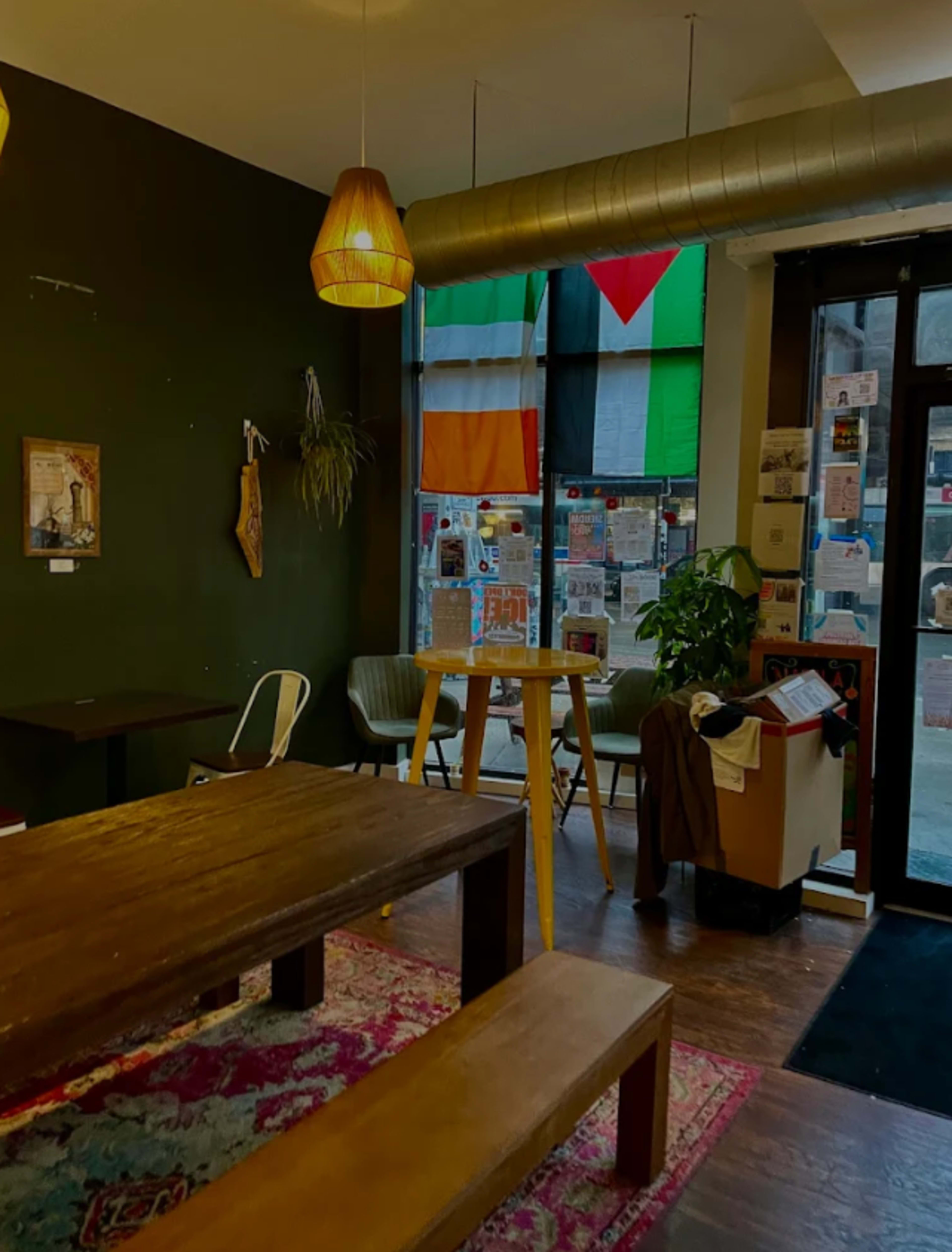 The interior of a café features wooden tables, hanging lights, and a corner with various flags and posters displayed in the window.