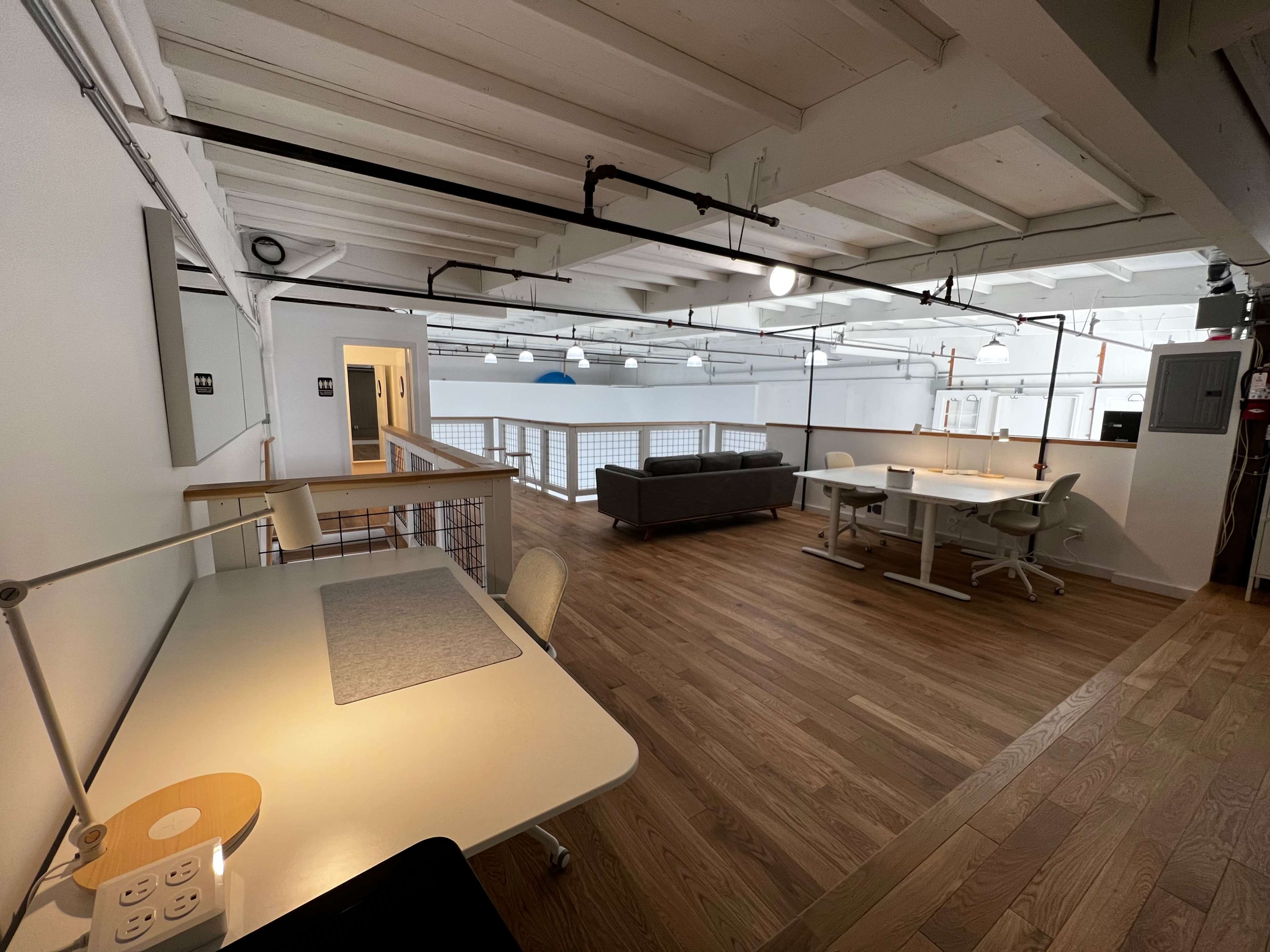 The image shows a spacious, modern office environment featuring desks, a couch, and wooden flooring.