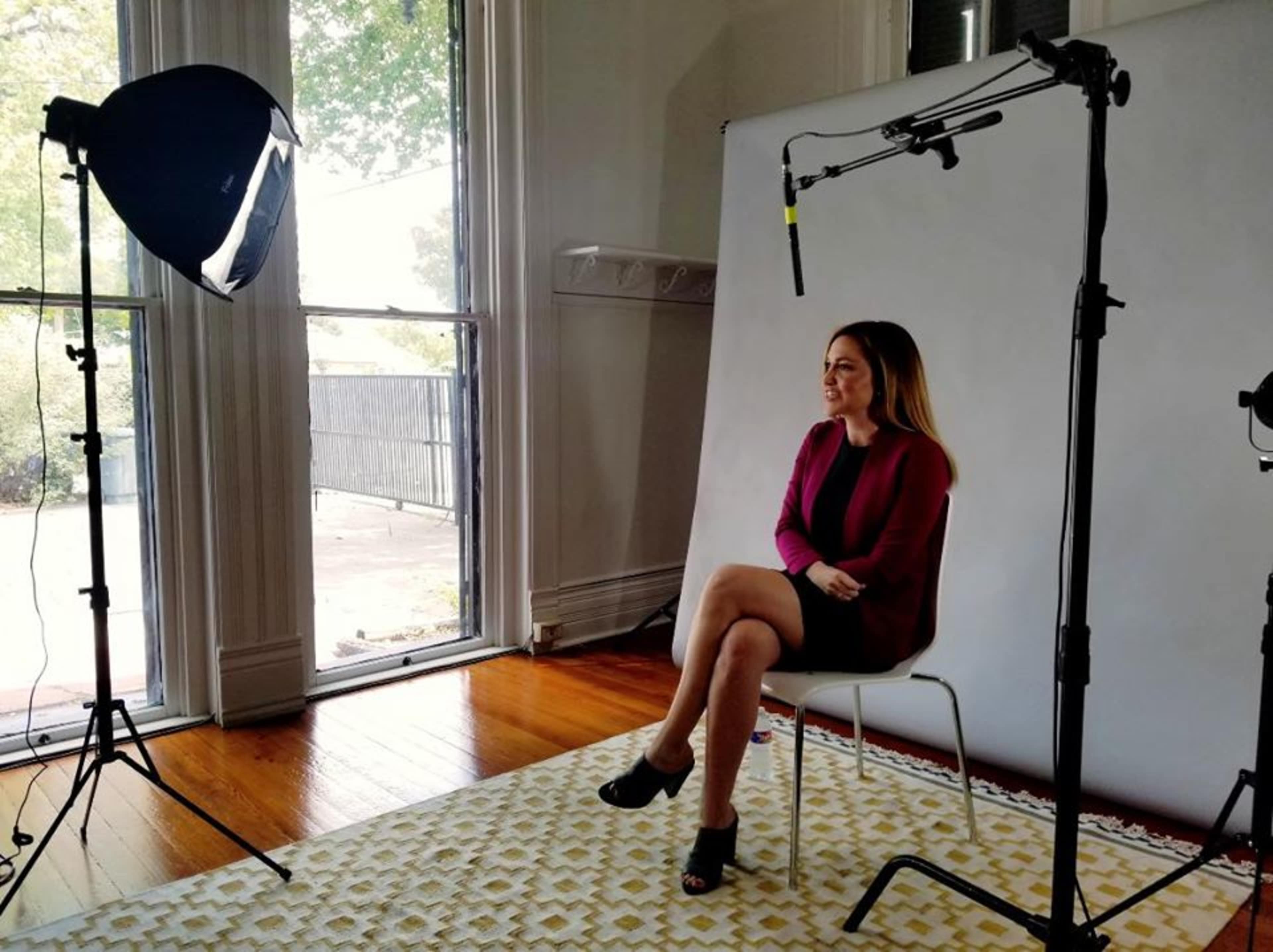 A woman sits on a chair in a brightly lit studio with a backdrop and professional lighting equipment.