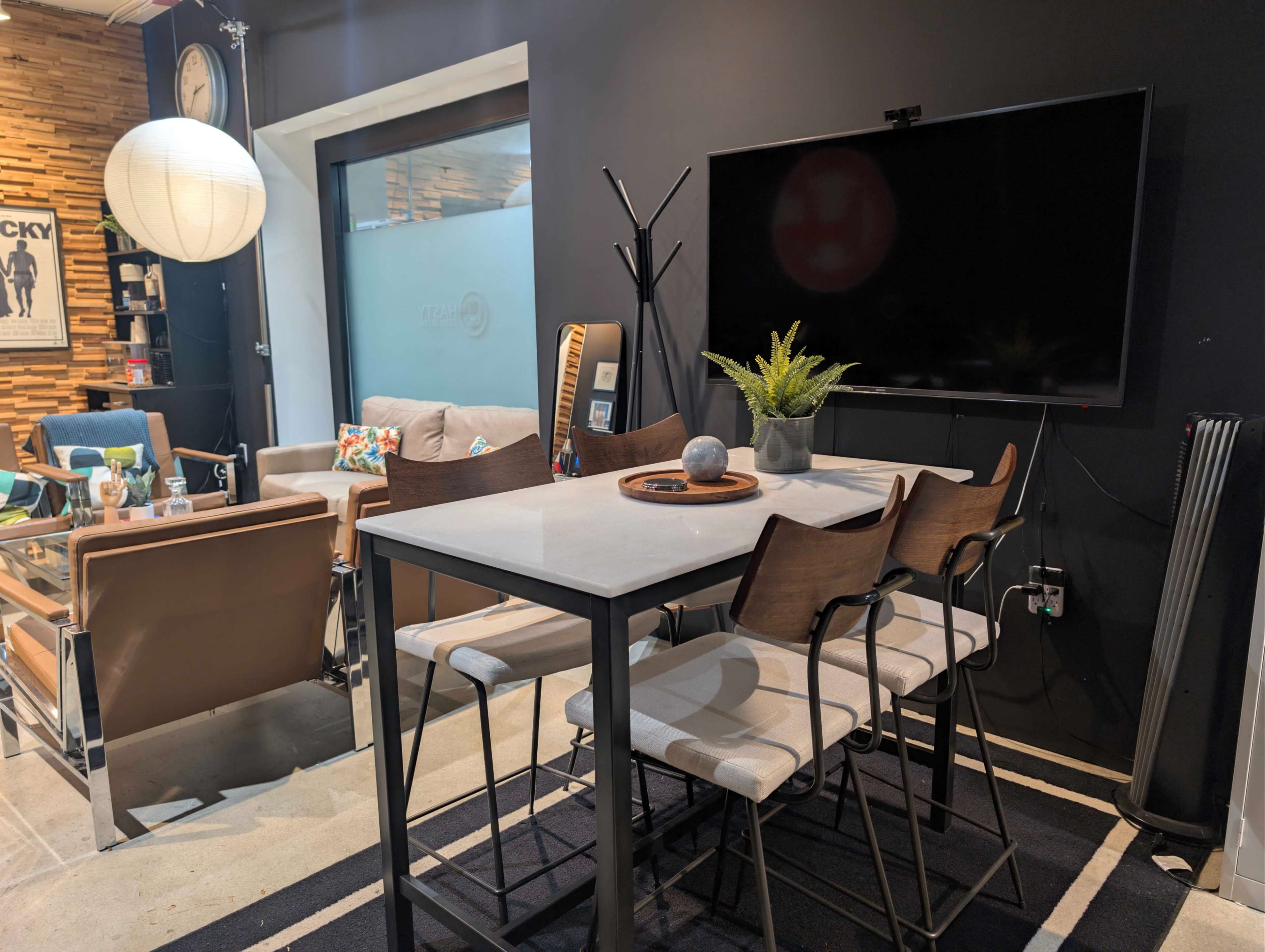The image shows a modern dining area with a rectangular table surrounded by chairs, a television mounted on the wall, and a seating area with a sofa and decorative elements in the background.