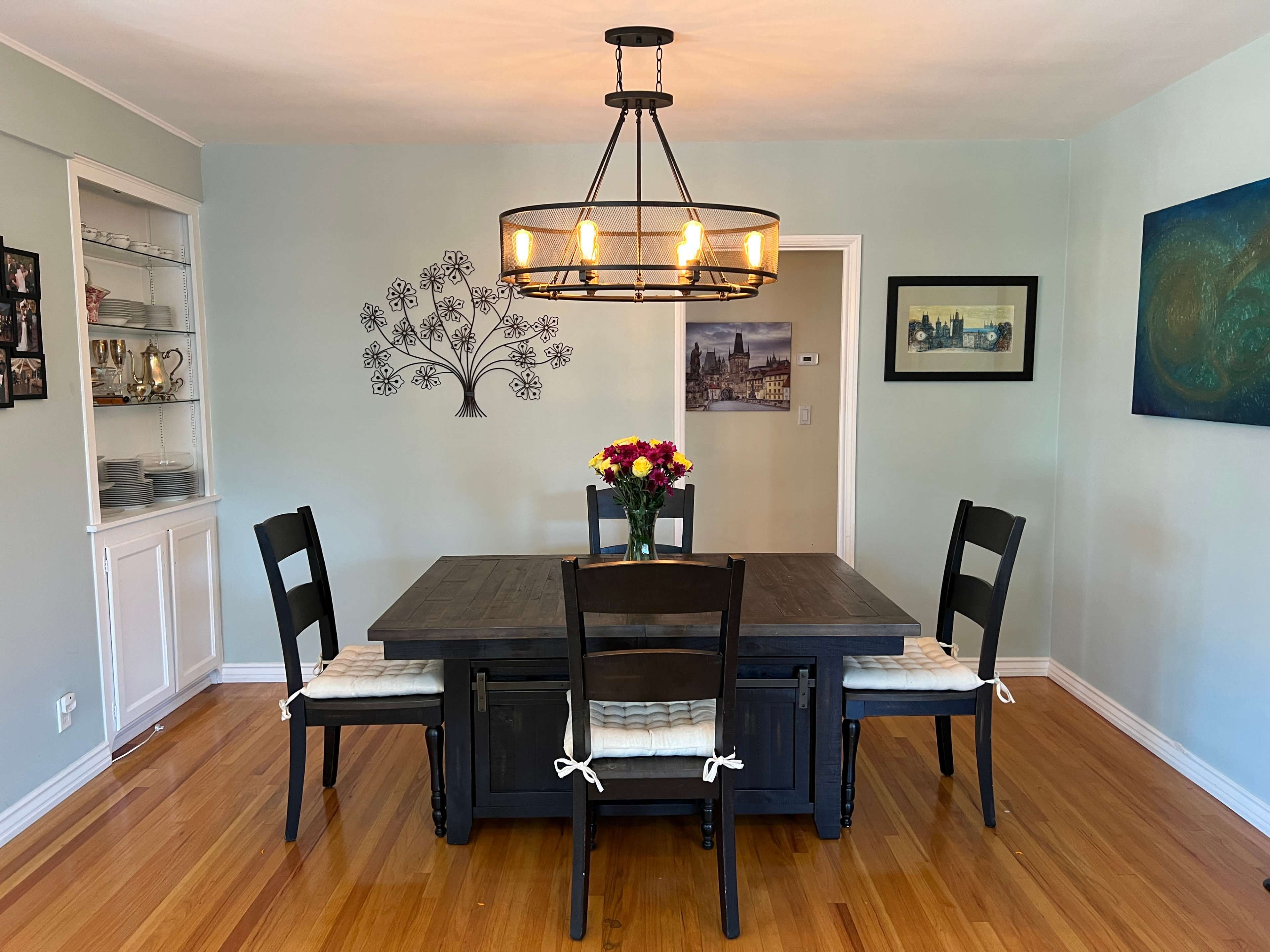 A dining room with a wooden table and chairs, a chandelier overhead, and a floral centerpiece on the table, accented by wall art and a light blue paint color.