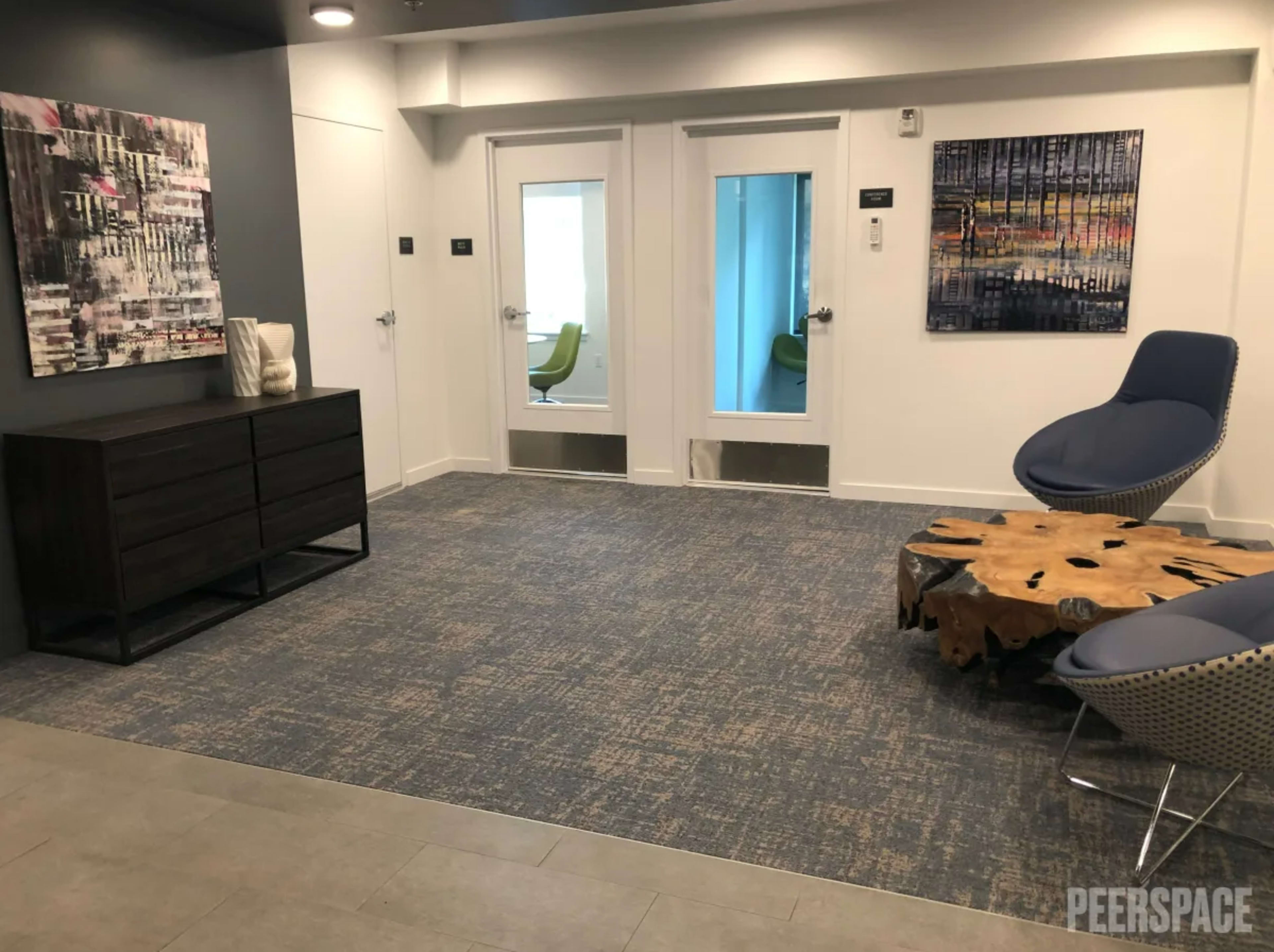 The image shows a modern lobby area with a wooden cabinet, a blue accent chair, and two glass doors leading to adjacent rooms.