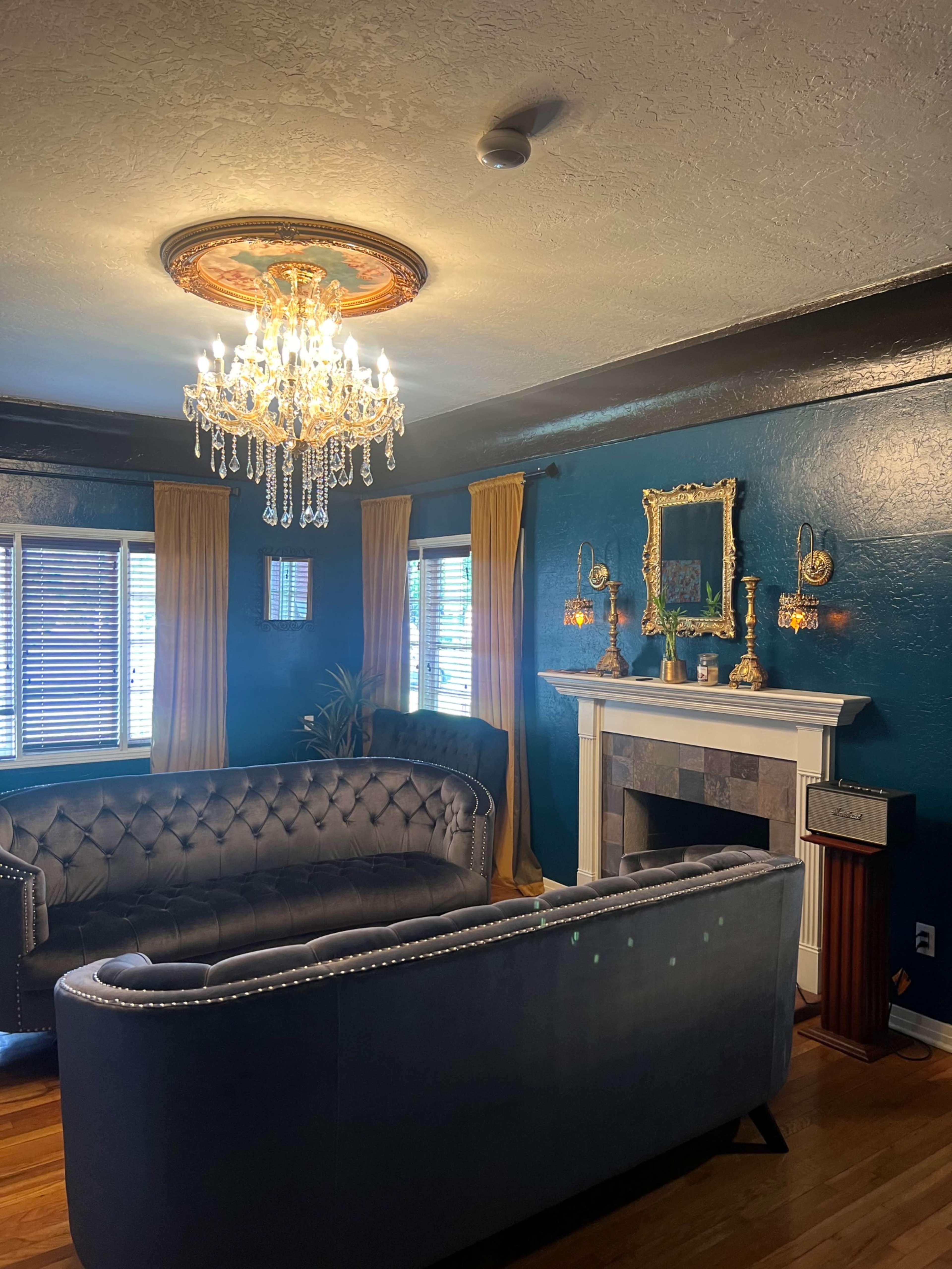 A living room with dark blue walls, a modern chandelier, plush sofas, and a fireplace framed by decorative mirrors and wall sconces.
