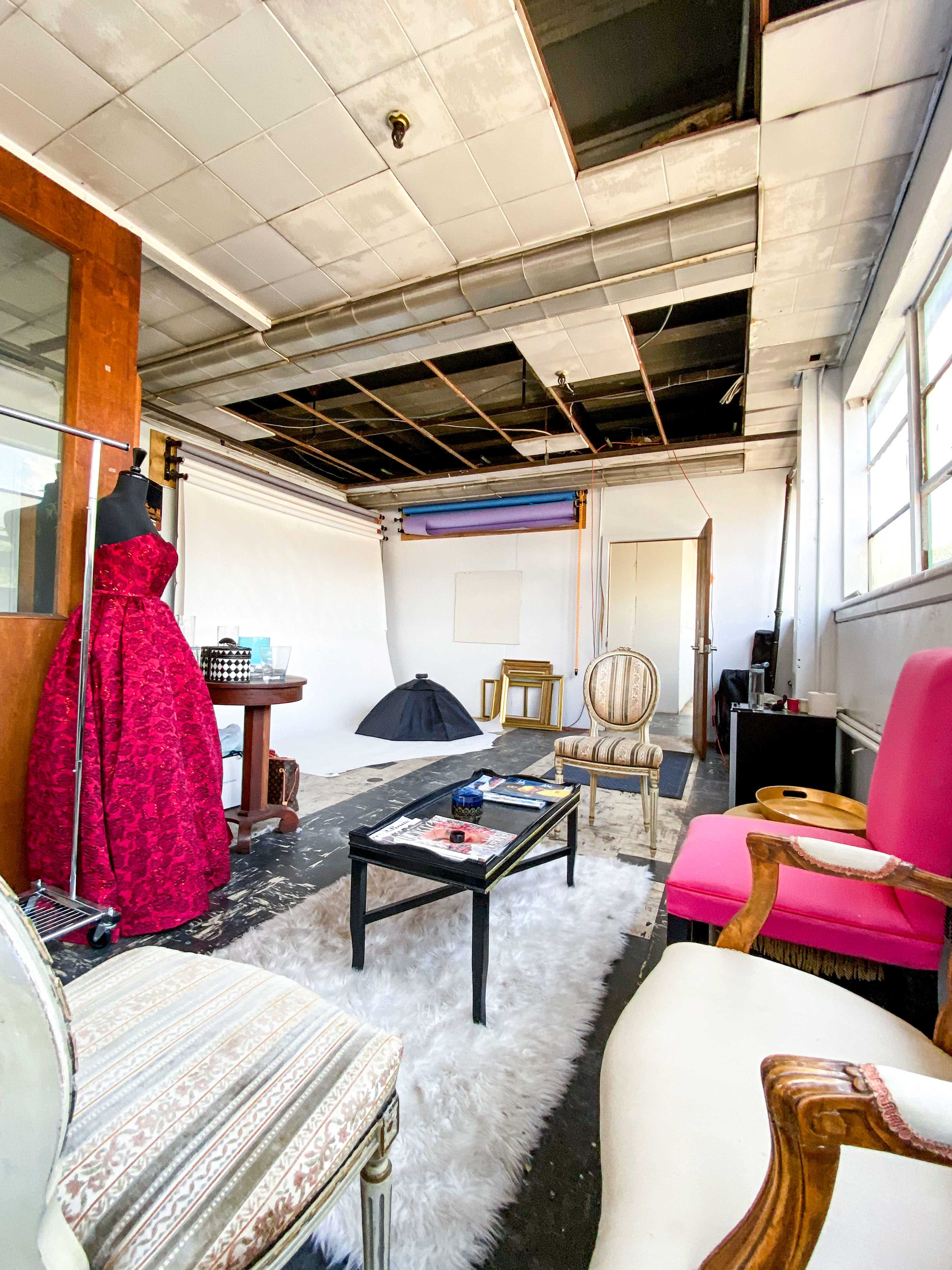 A spacious studio with exposed ceiling tiles, a large pink chair, a vintage table with a mannequin dressed in a red gown, and various props arranged around a white backdrop.