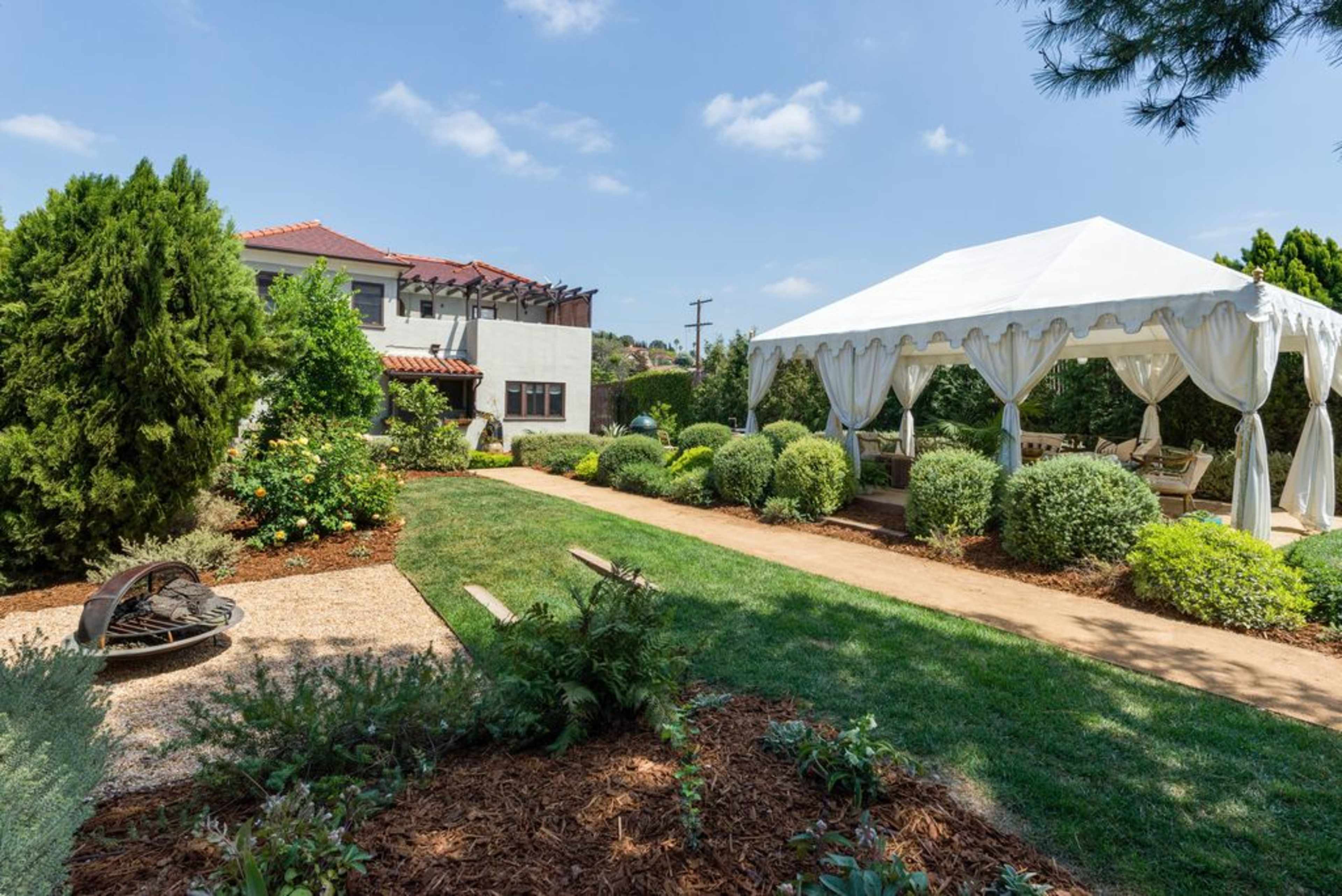A well-maintained garden features a white tent on a grassy area surrounded by neatly trimmed hedges and plants, with a house visible in the background.