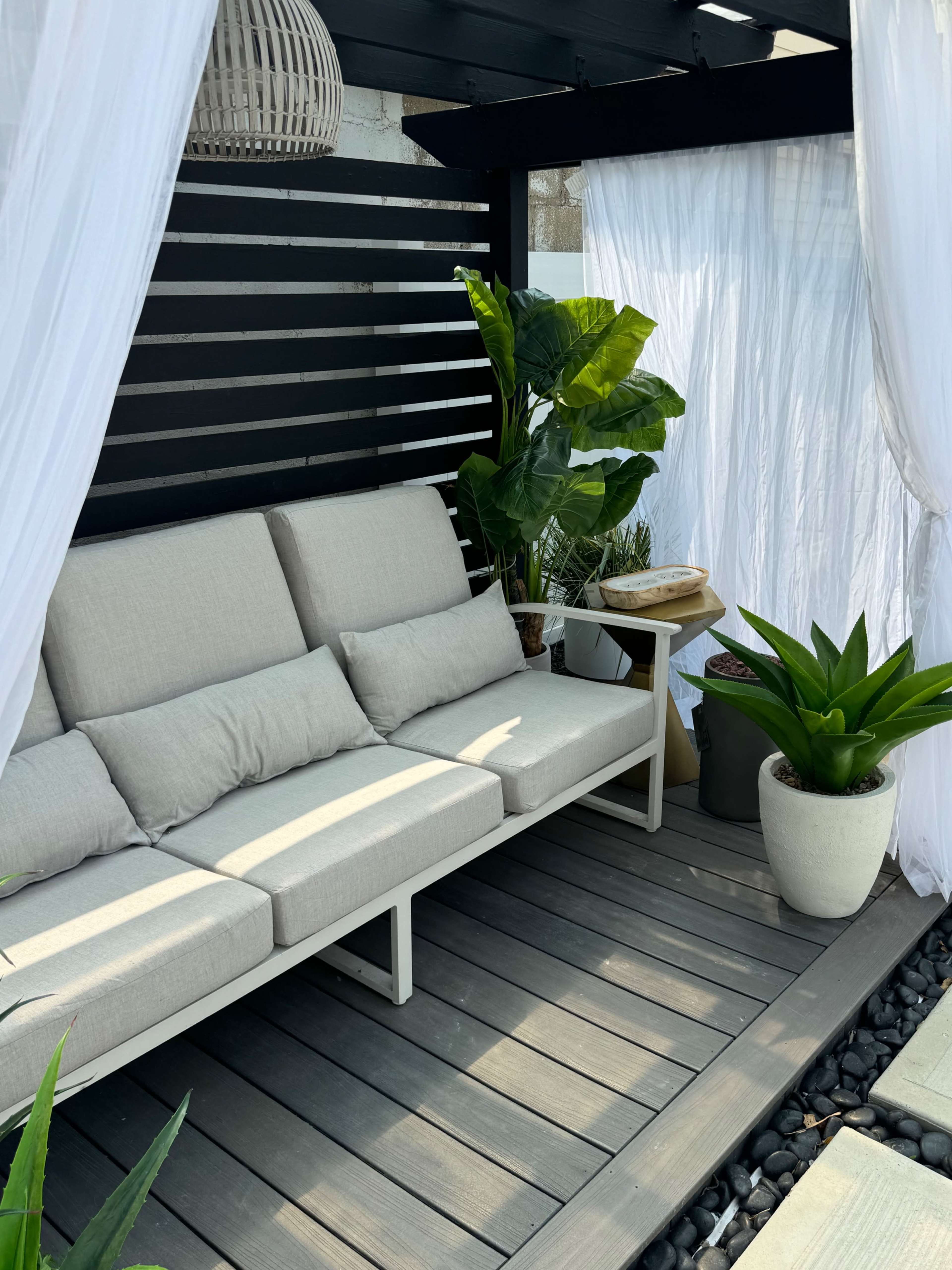 The image shows a cozy outdoor seating area with a beige sofa, potted plants, and sheer white curtains within a black pergola.