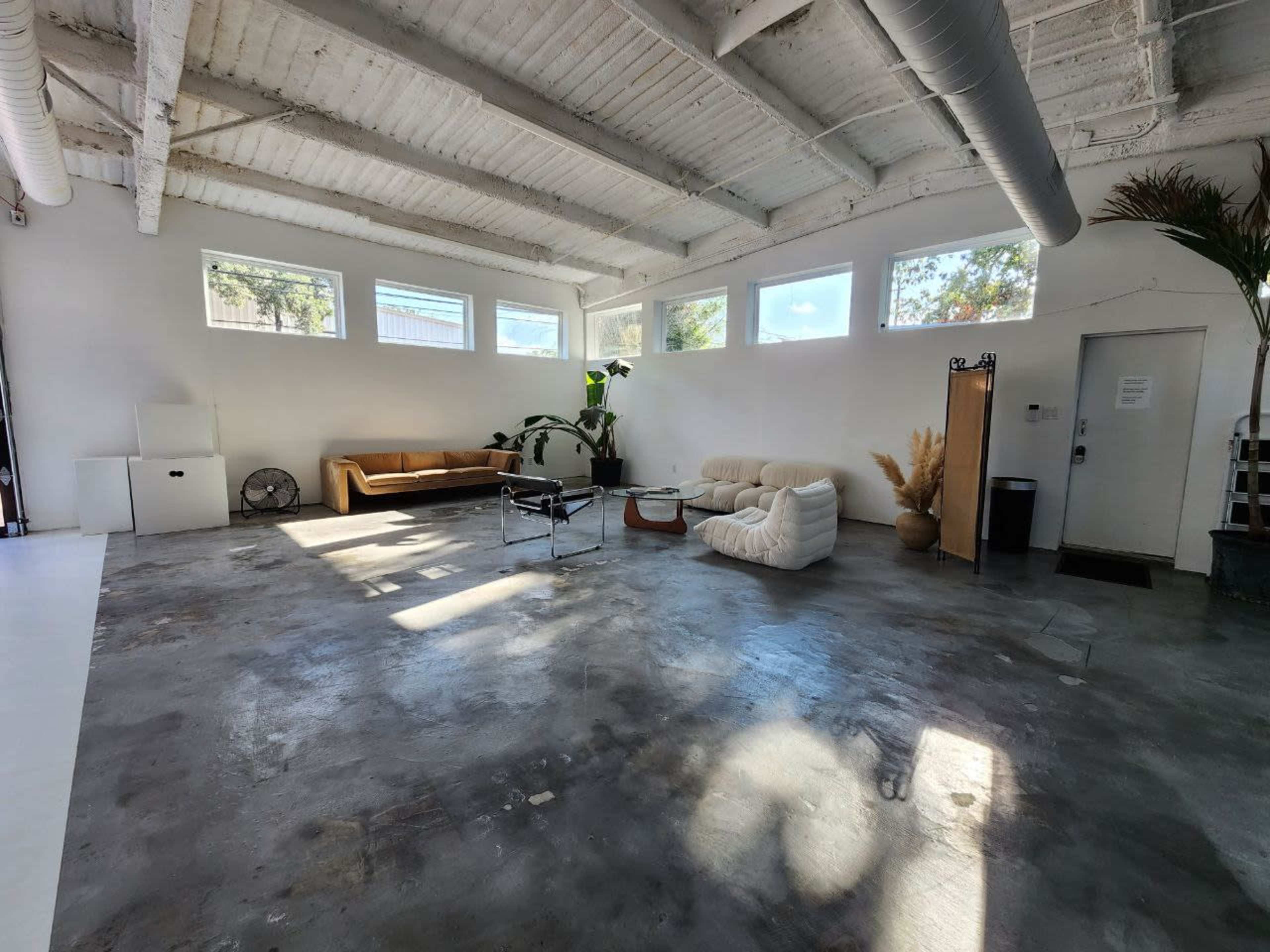 A spacious, minimalist room with a concrete floor, large windows, and several pieces of furniture including a couch, armchair, and a small table.