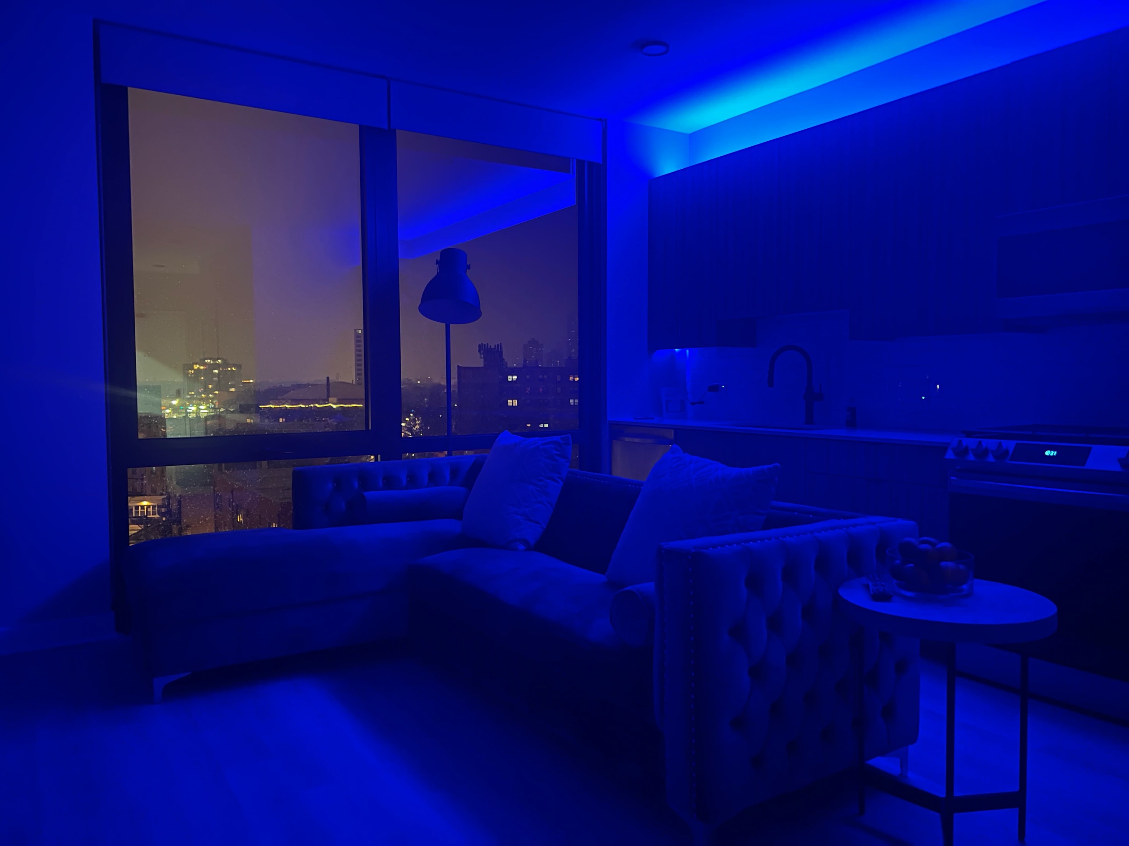 A dimly lit modern apartment features a blue-tinted sofa, a small table with fruit, and a view of a city skyline through large windows.