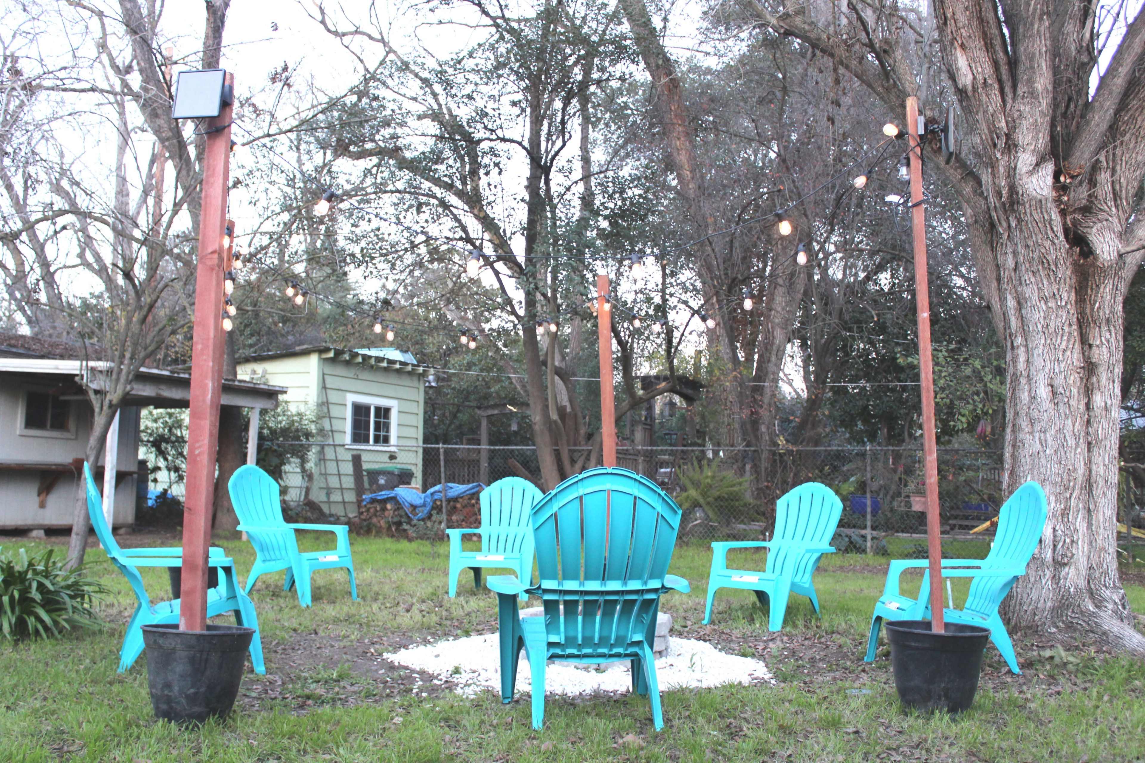 A circular arrangement of turquoise Adirondack chairs surrounds a fire pit in a backyard illuminated by string lights hung between wooden posts.