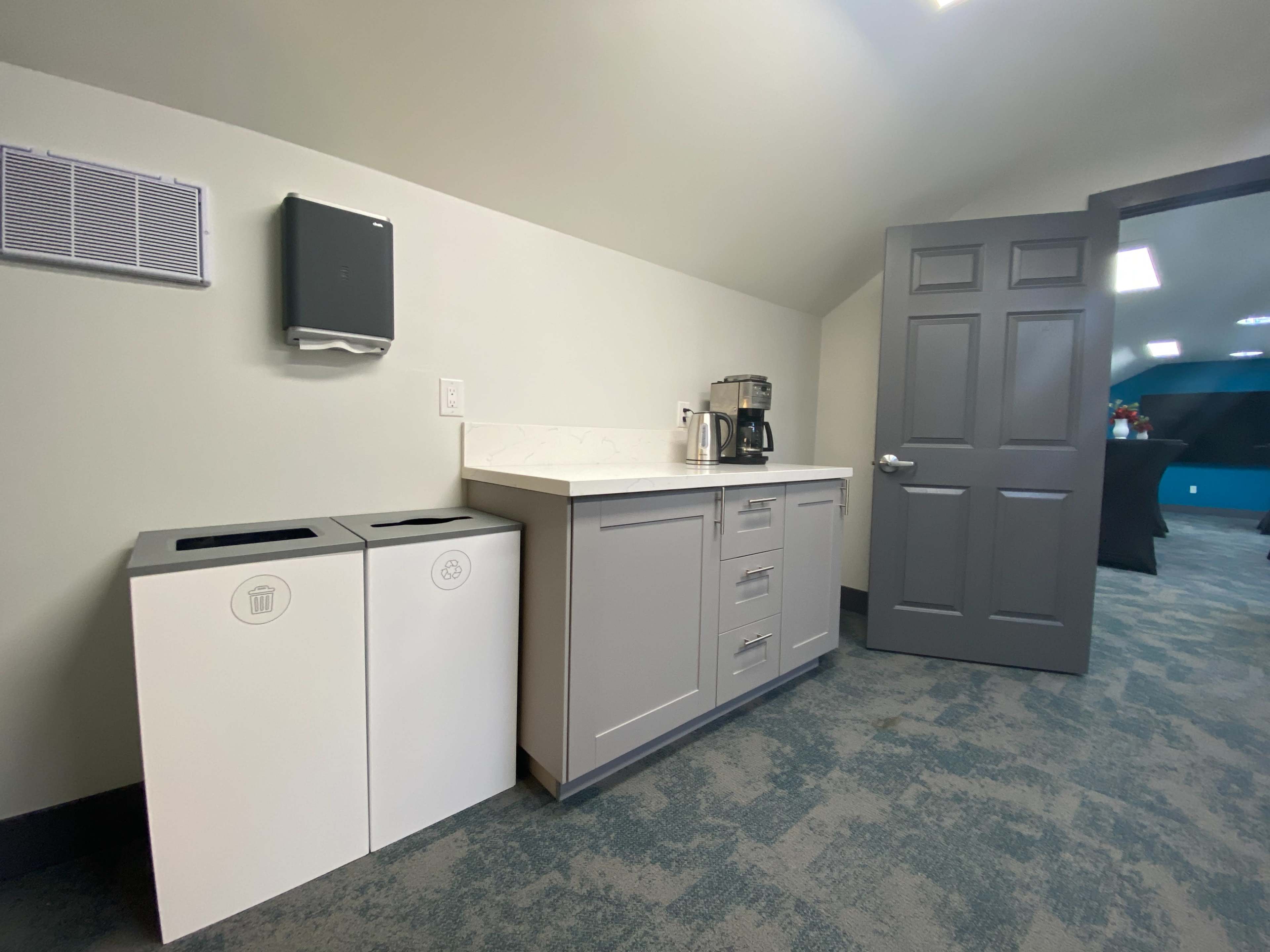 The image shows a small kitchenette area featuring a countertop with a coffee maker and cabinets, alongside two bins for recycling and trash.