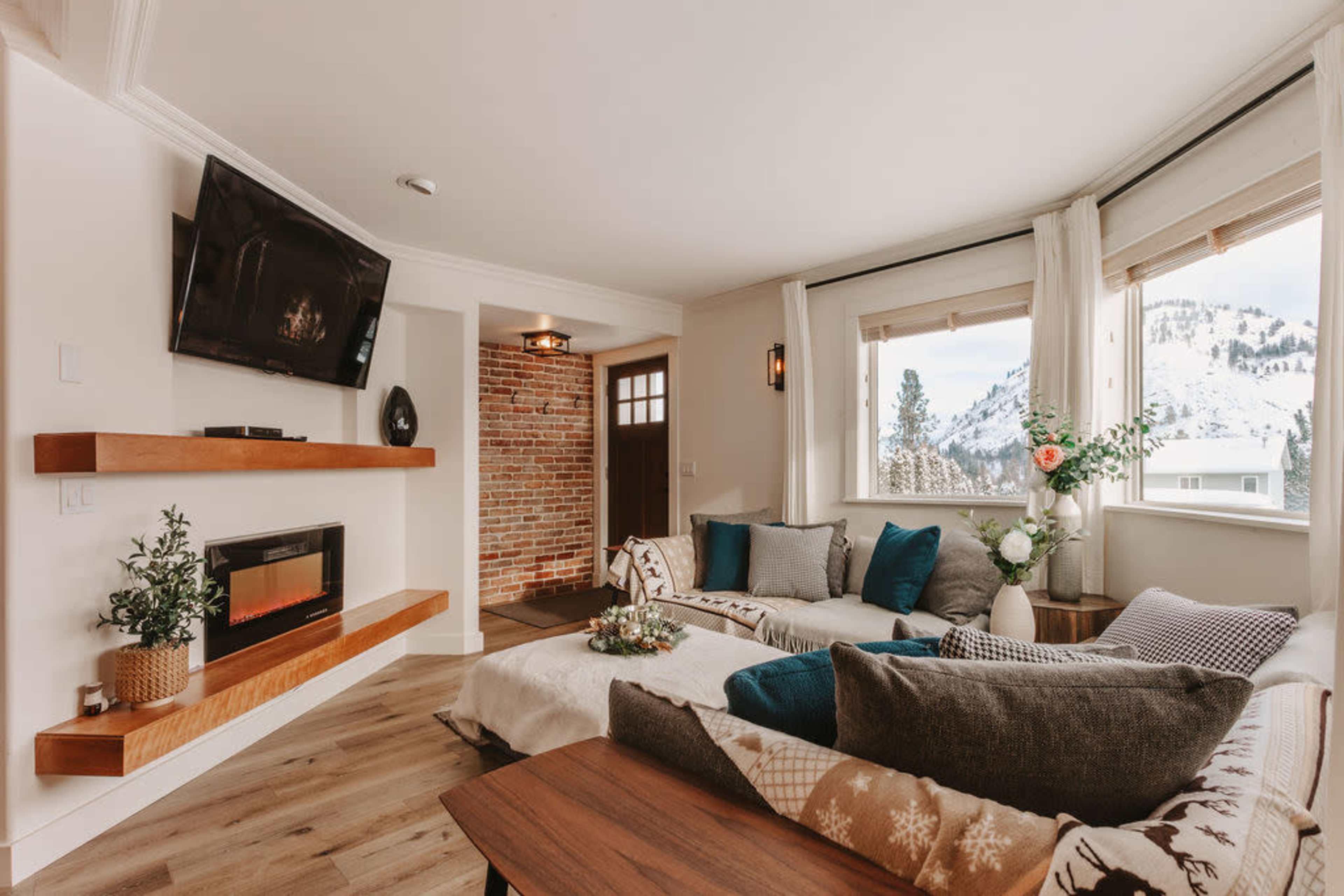 The living room features a modern design with a sectional sofa, a mounted television, a wall of exposed brick, and large windows overlooking a snowy landscape.