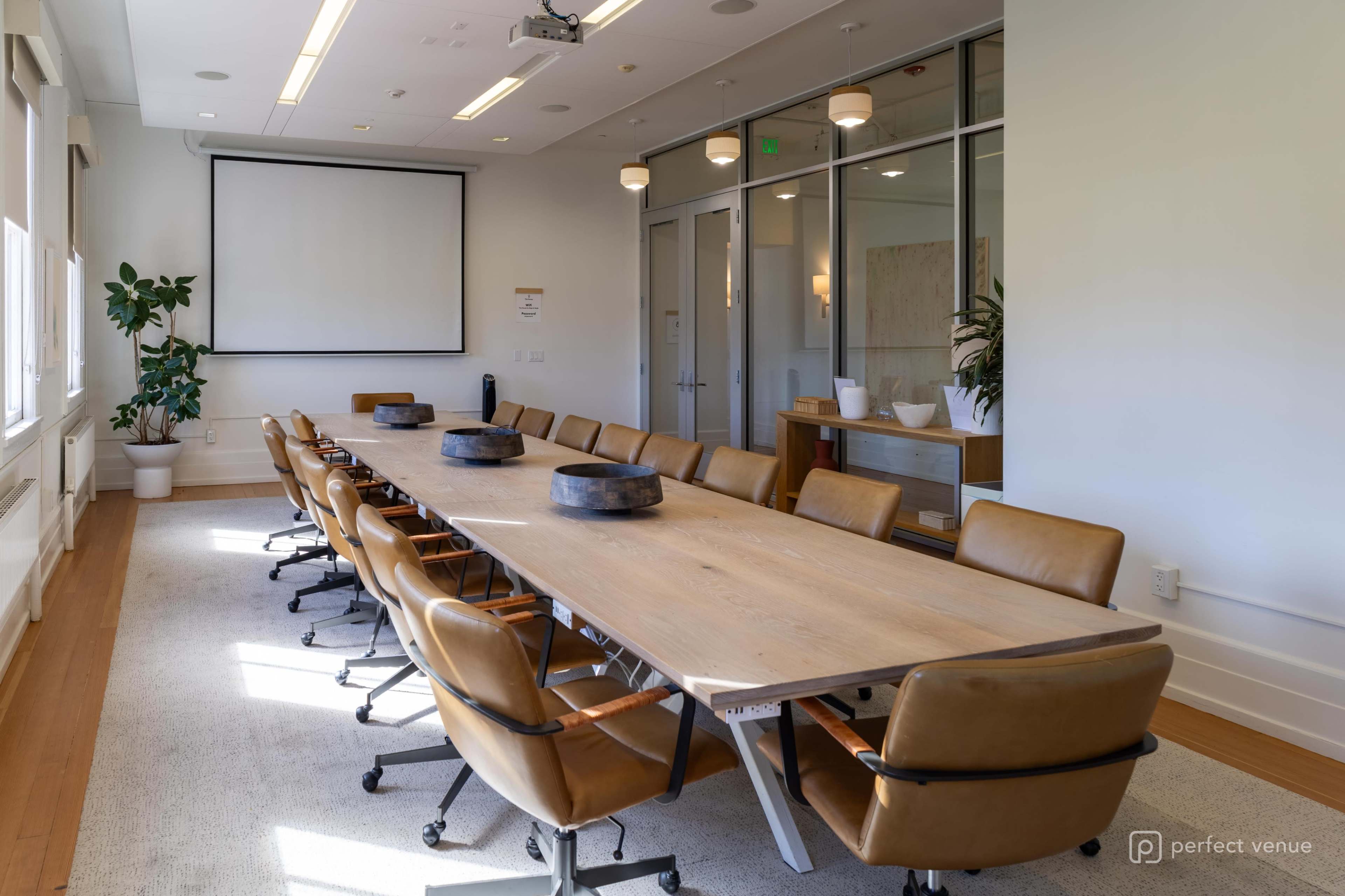 A large conference room features a long wooden table surrounded by brown leather chairs, with a projector screen and glass doors in the background.