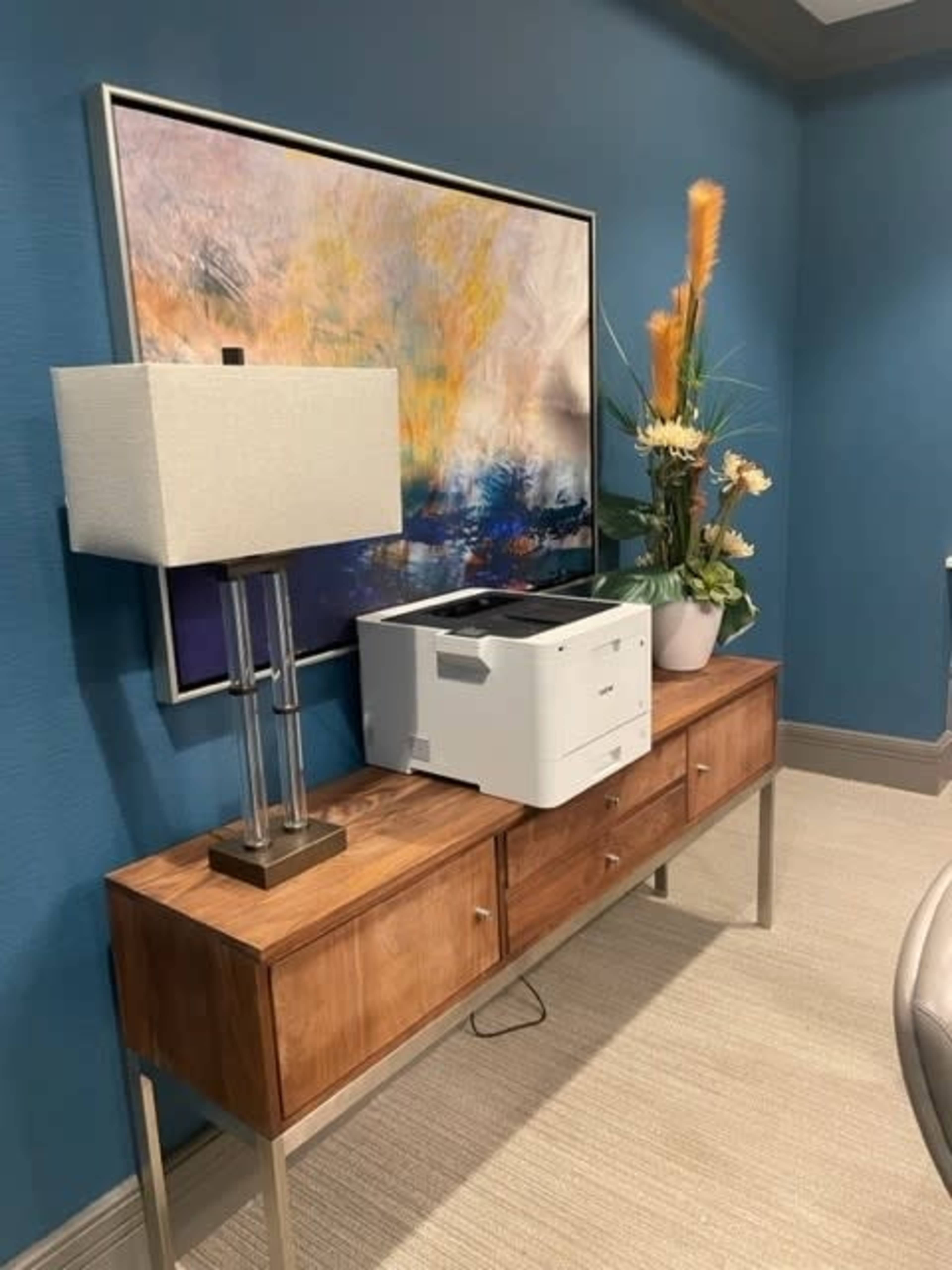 A printer sits on a wooden console table beneath a decorative lamp and a vibrant wall painting, accompanied by a floral arrangement.