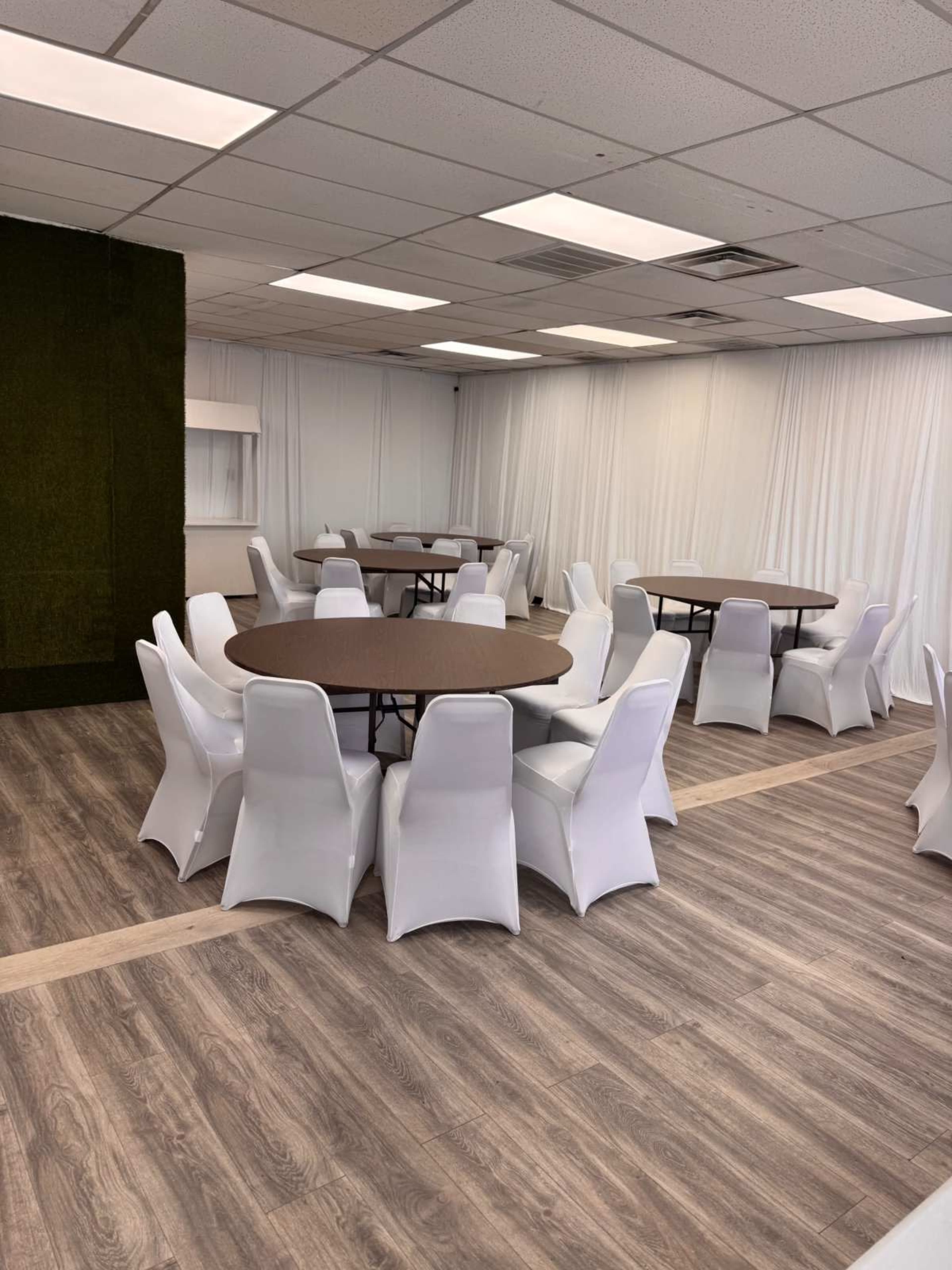 The image shows a spacious room with several round tables surrounded by white chairs, with a light wood floor and white drapes.