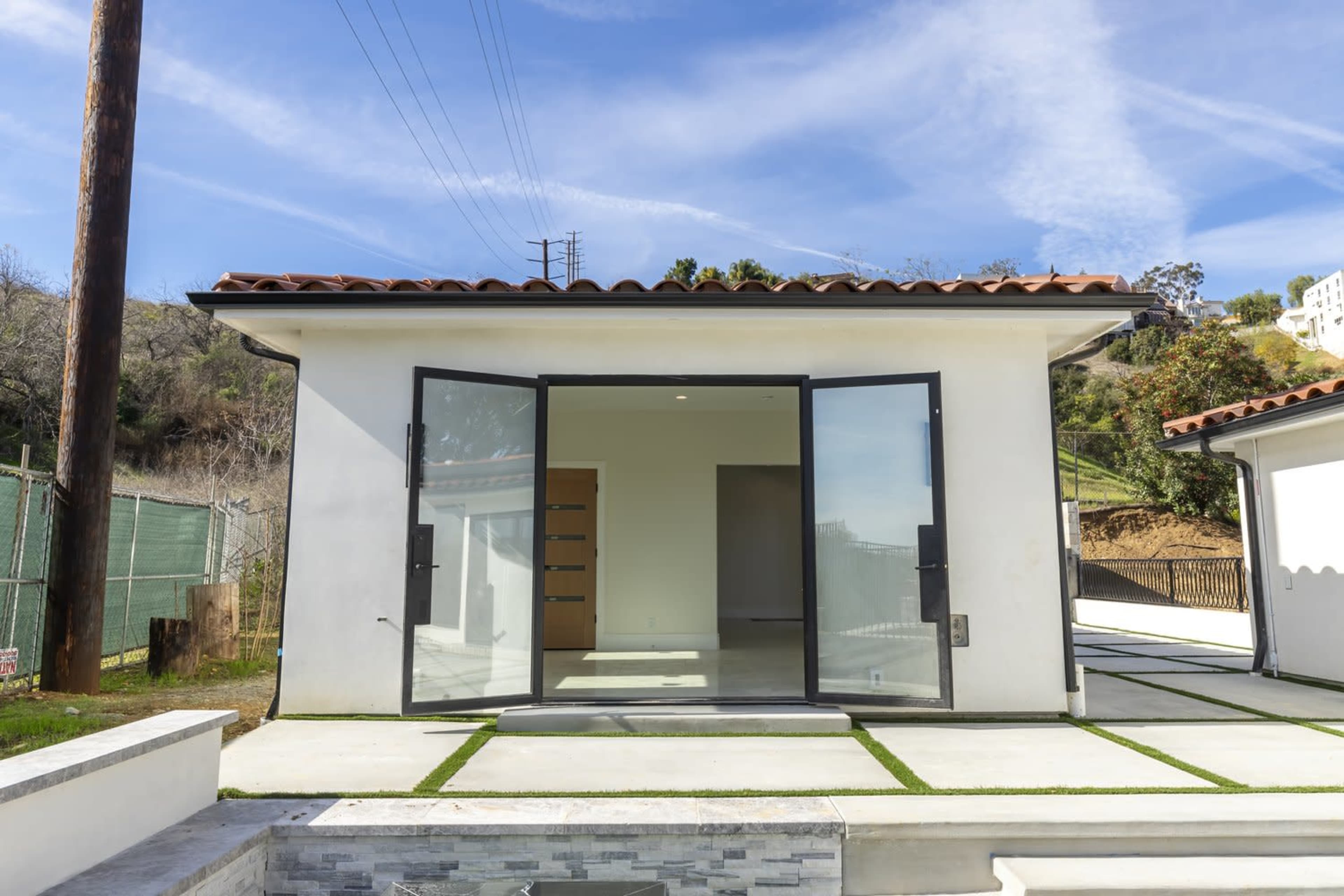 The image shows a modern, single-story house with large glass doors opening onto a concrete patio, surrounded by greenery and distant hills.