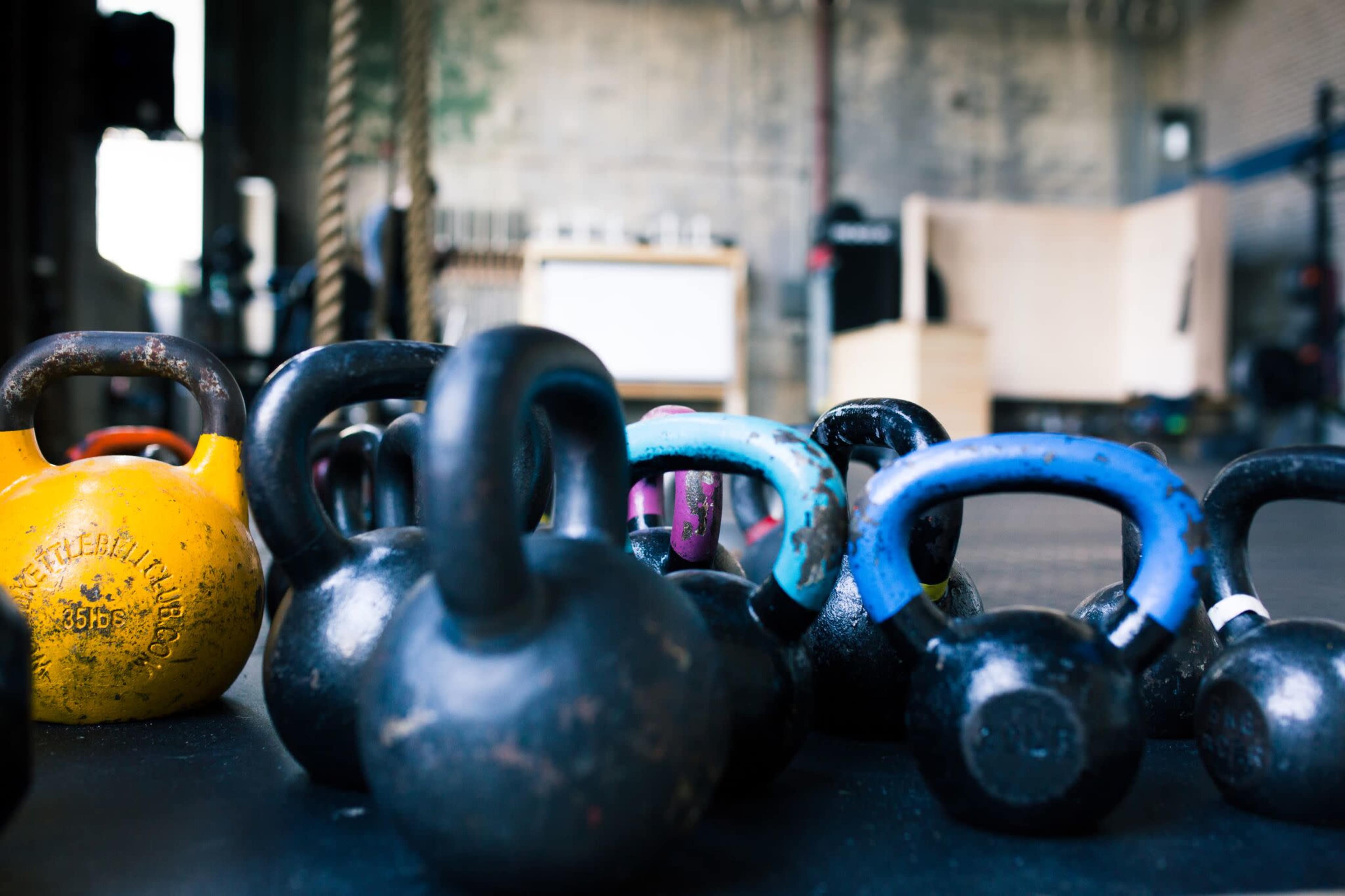 A group of kettlebells in various colors and sizes is arranged on a gym floor.