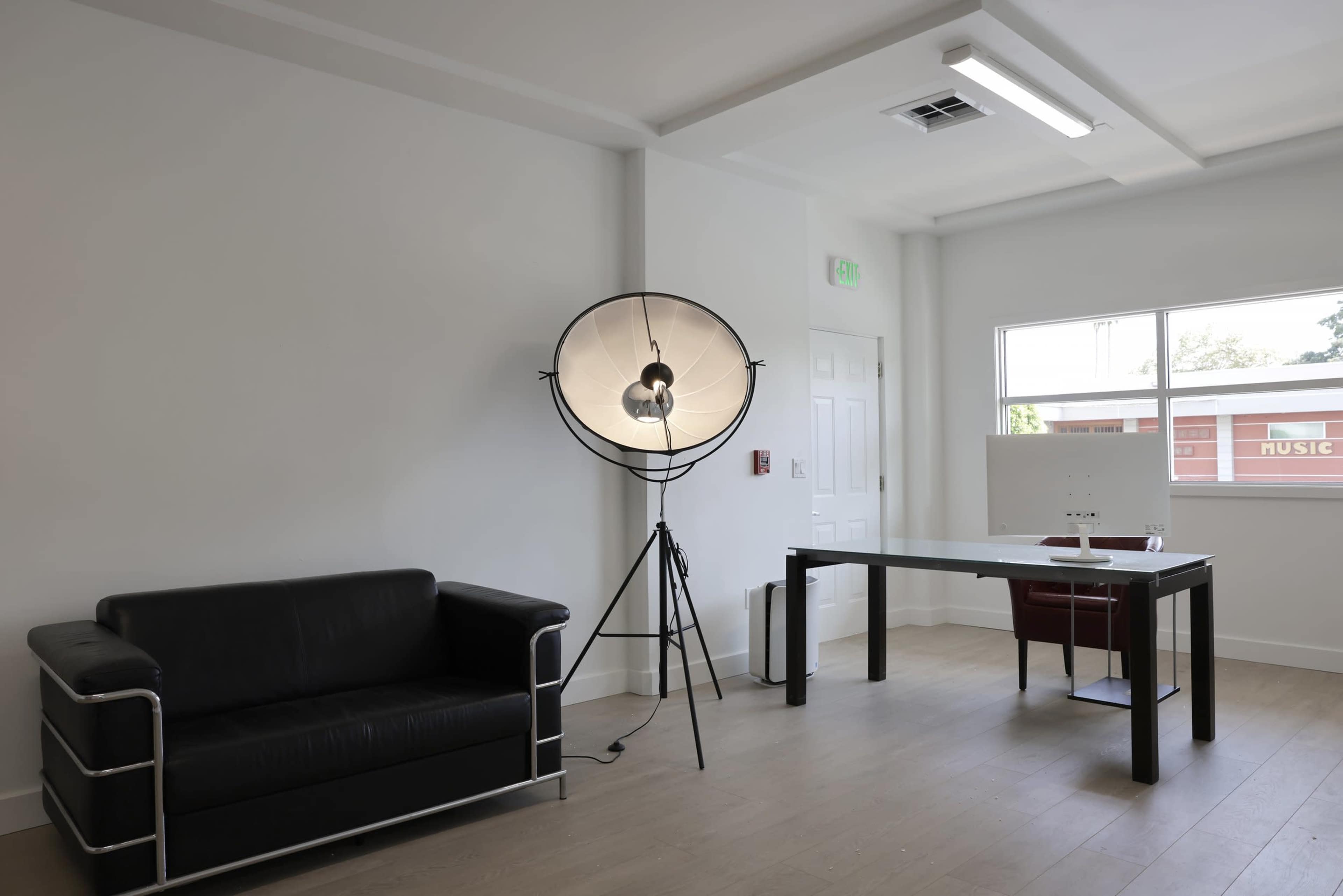The image shows a minimalist office space with a black leather couch, a desk with a chair, and a large floor lamp.