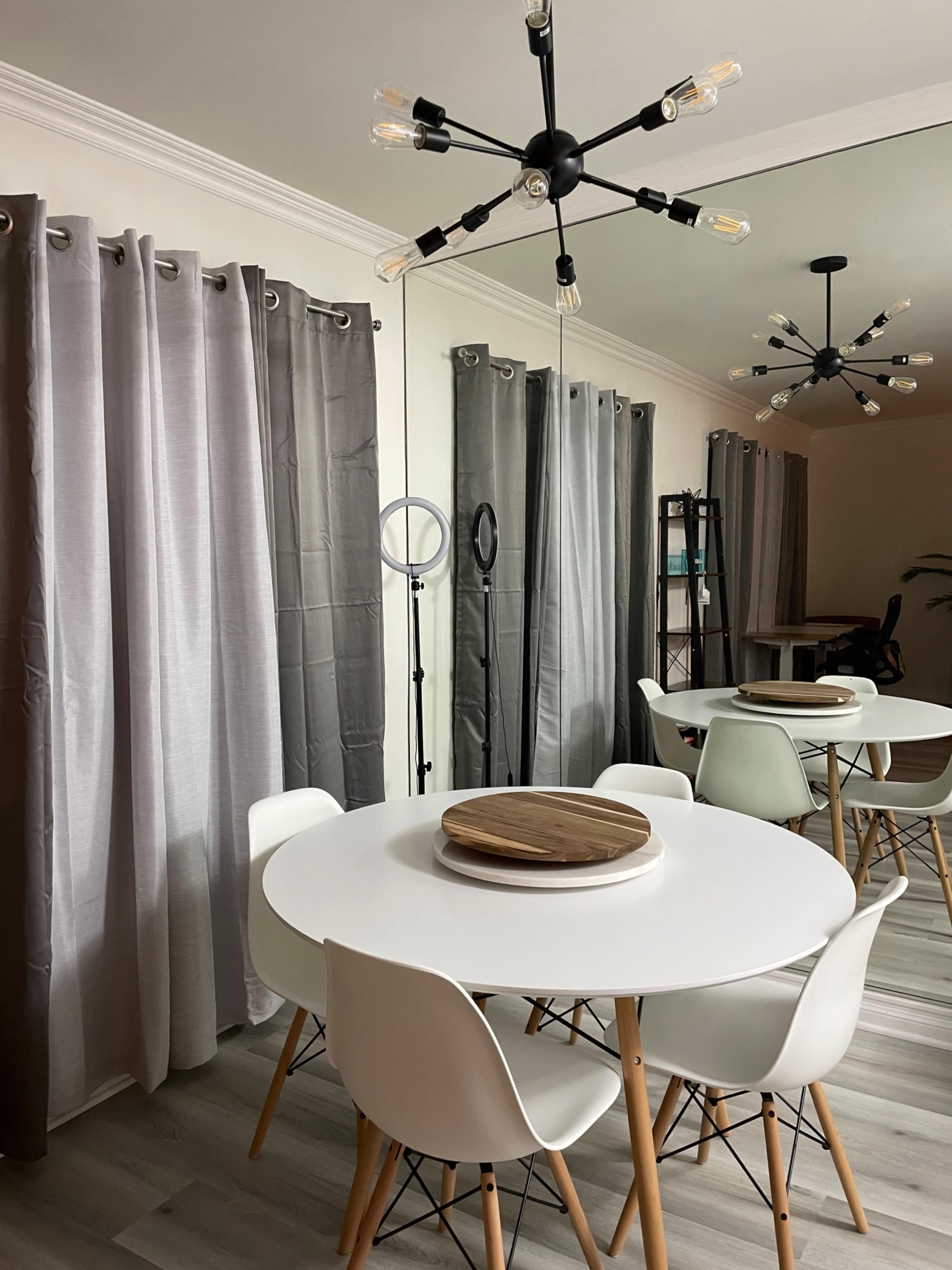 A modern dining area features a round white table surrounded by white chairs, with gray curtains and a geometric light fixture overhead.