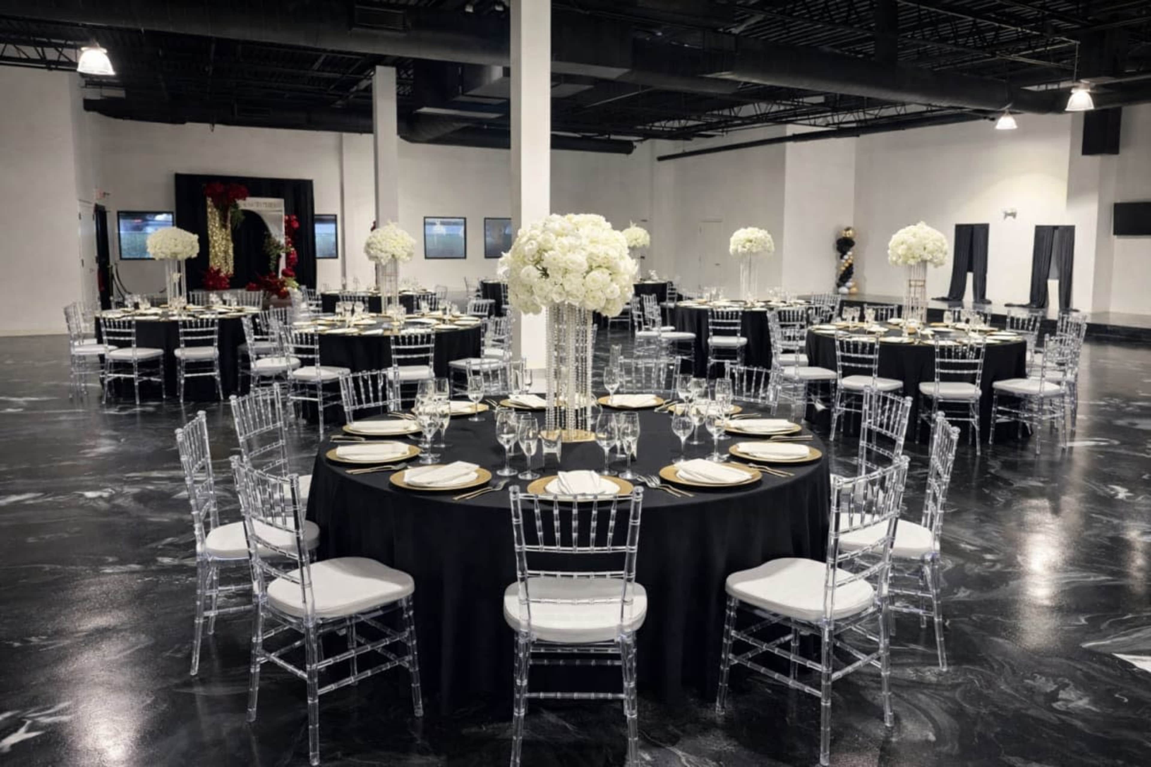 The image shows a spacious event venue set up with several round tables covered in black tablecloths, adorned with white floral centerpieces, and surrounded by clear acrylic chairs.