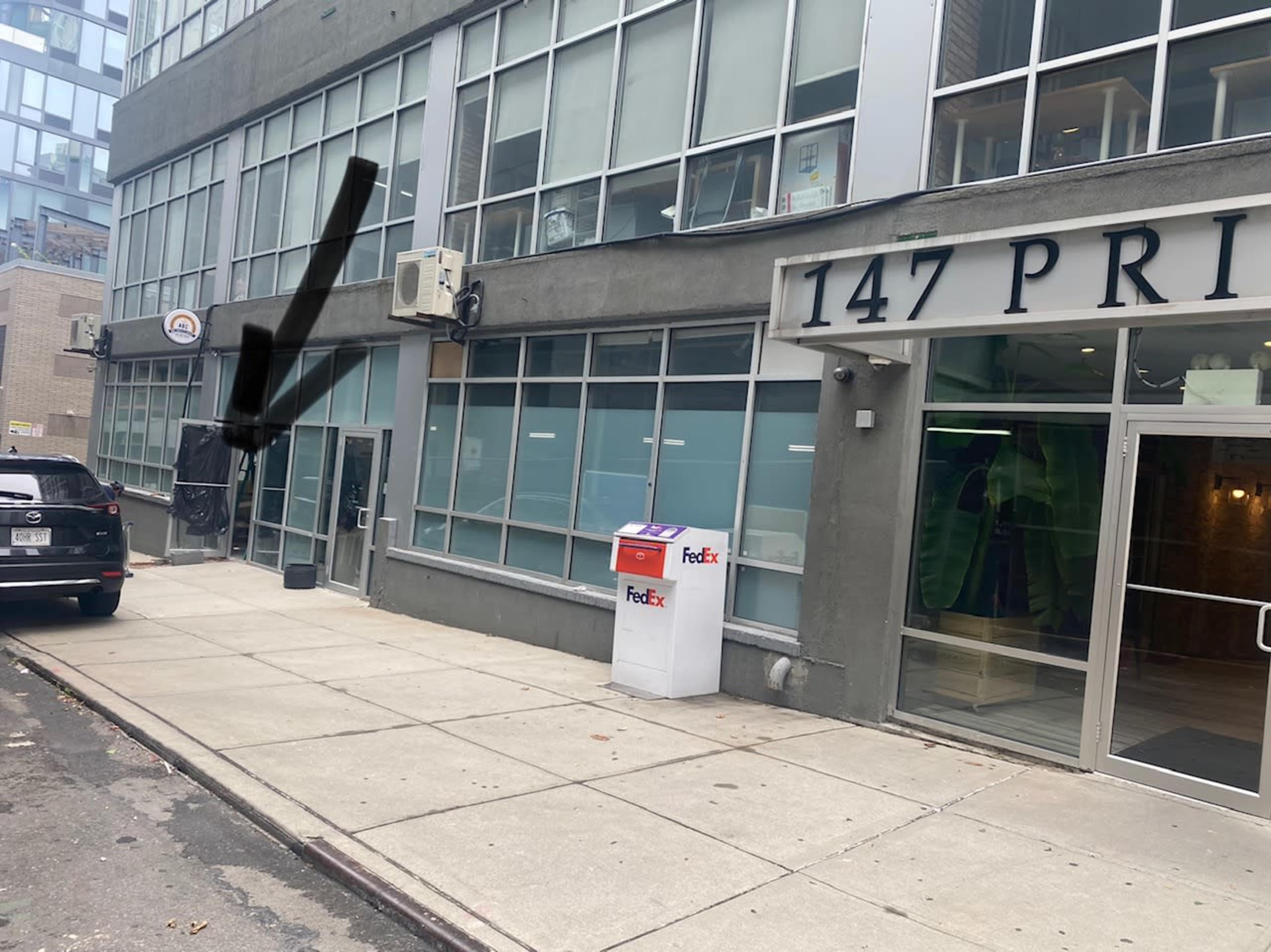 A sidewalk in front of a modern building with large windows, featuring a FedEx drop box and a dark awning above the entrance.