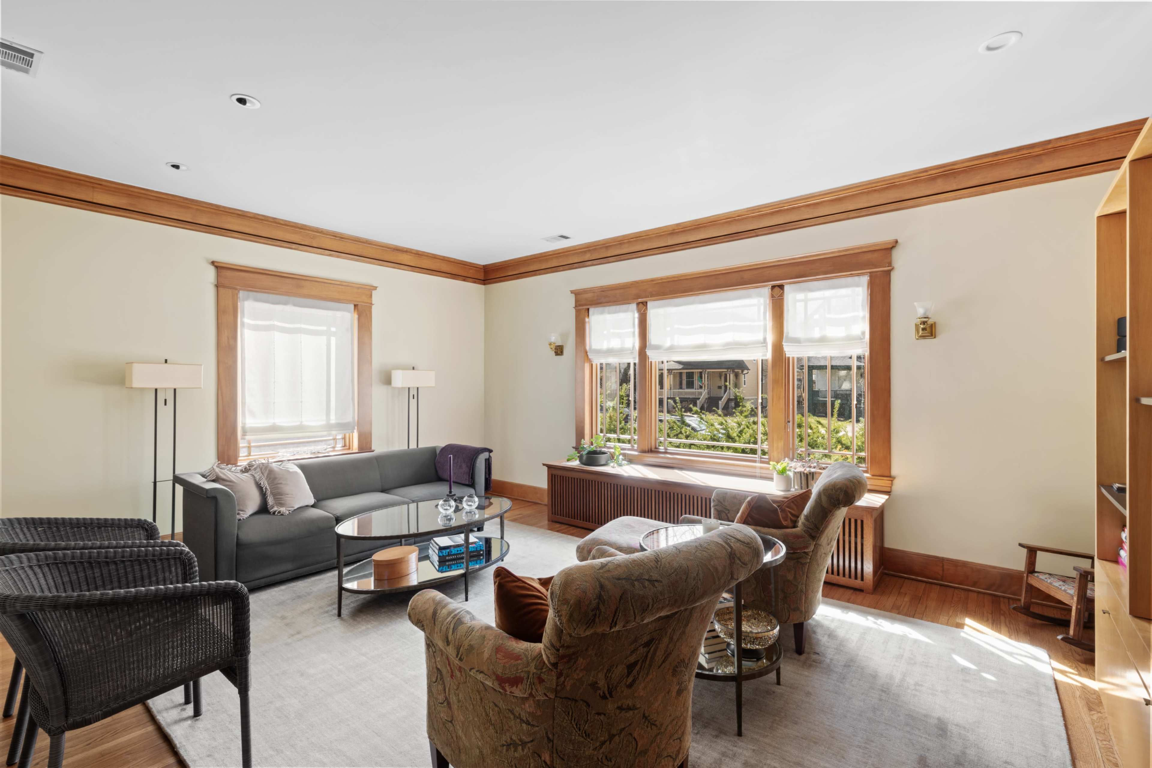 A cozy living room features a gray couch, two patterned armchairs, a glass coffee table, and large windows with light curtains.