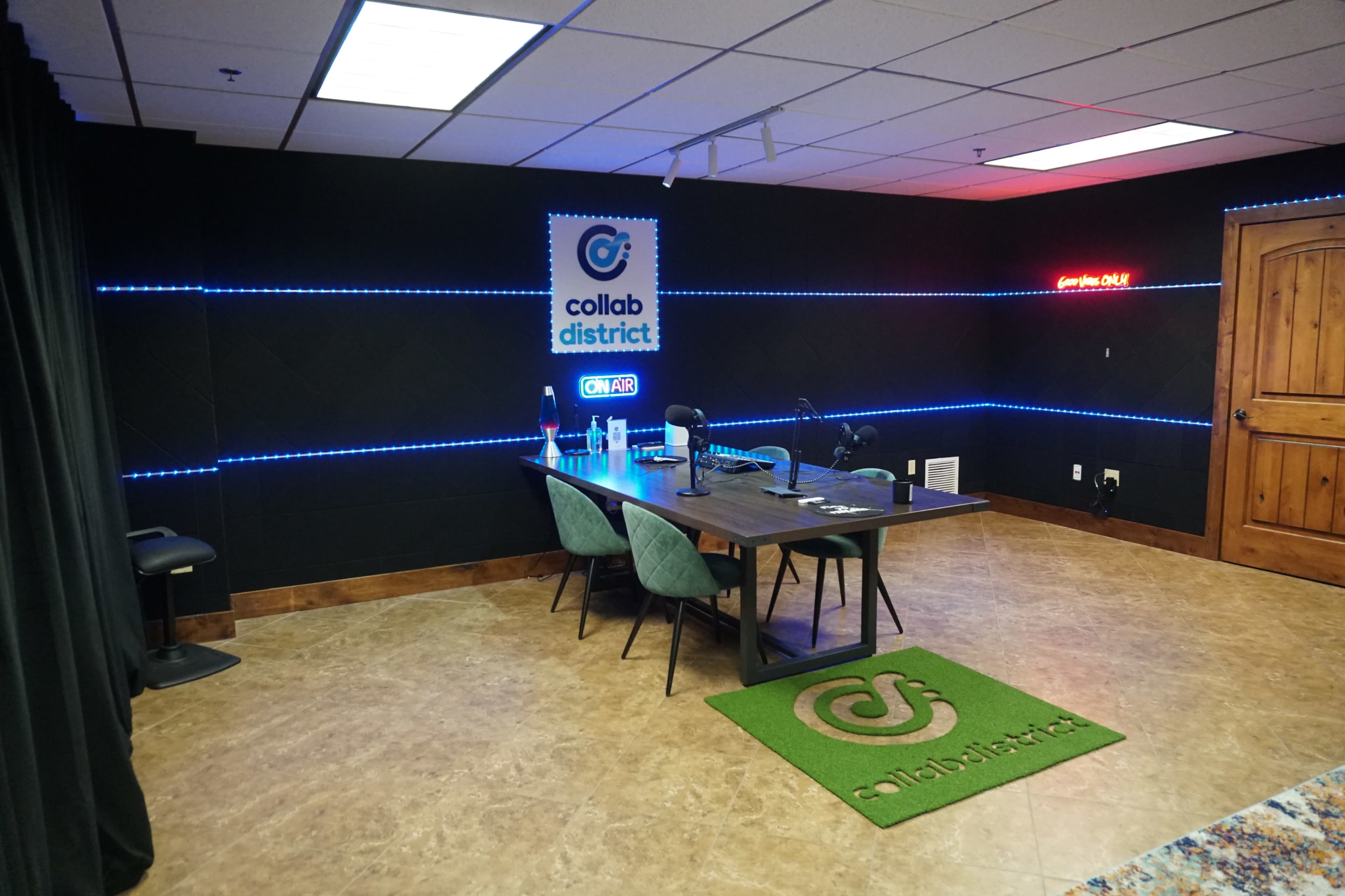 A modern podcasting studio with a wooden table, green chairs, illuminated walls, and a sign that reads "collab district ON AIR."