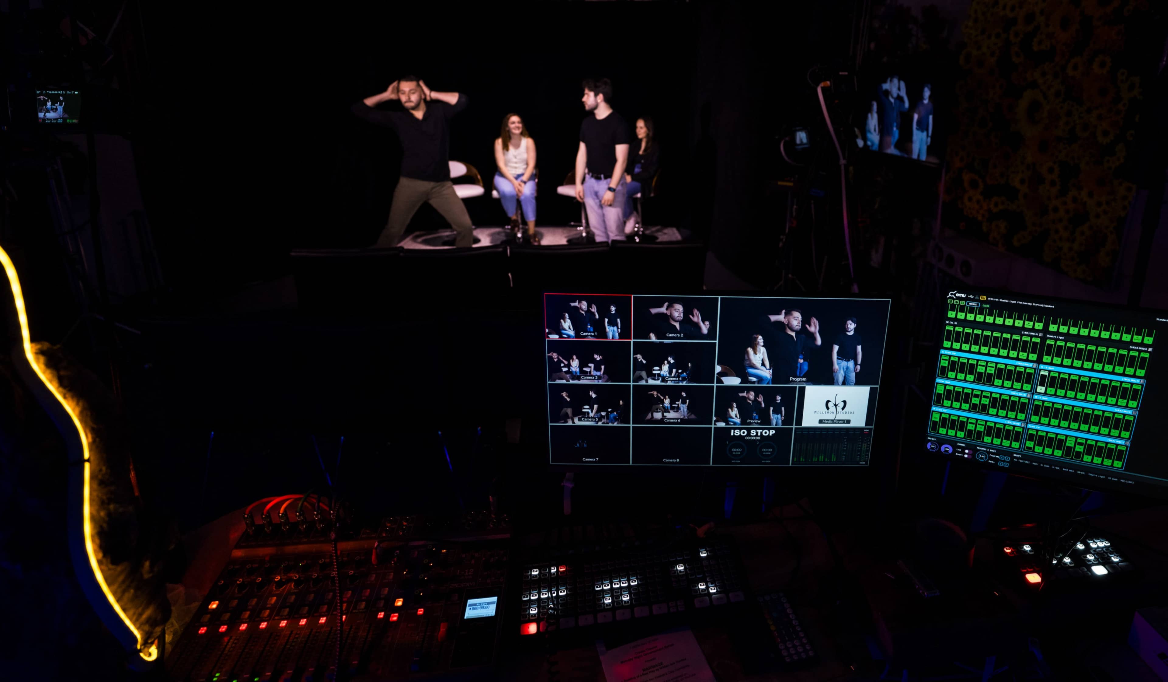 A production control room displays multiple camera feeds while a live performance occurs on stage in the background.