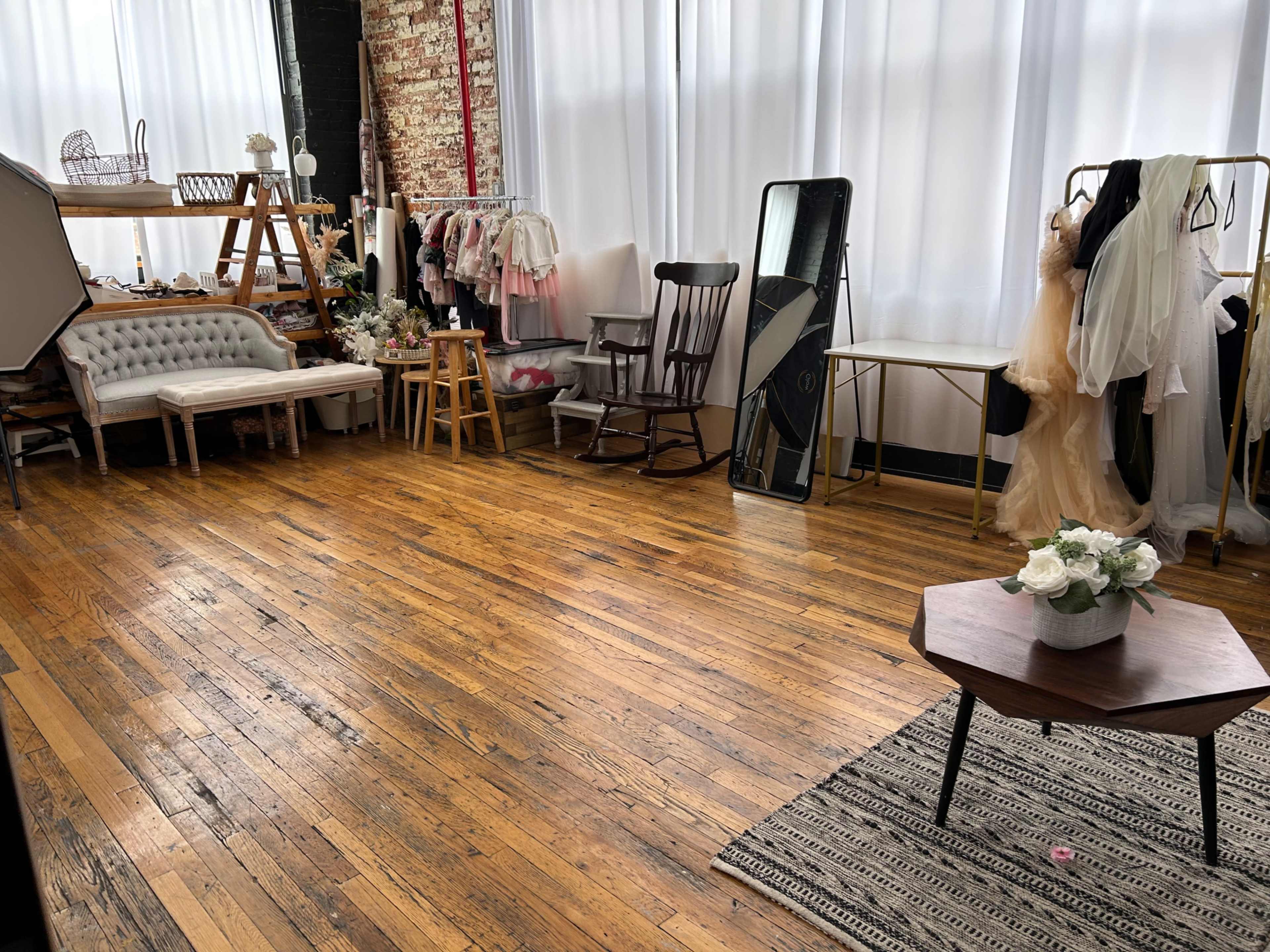 A spacious studio with wooden floors, large windows, and various clothing racks and furniture, including a rocking chair and a coffee table with flowers.