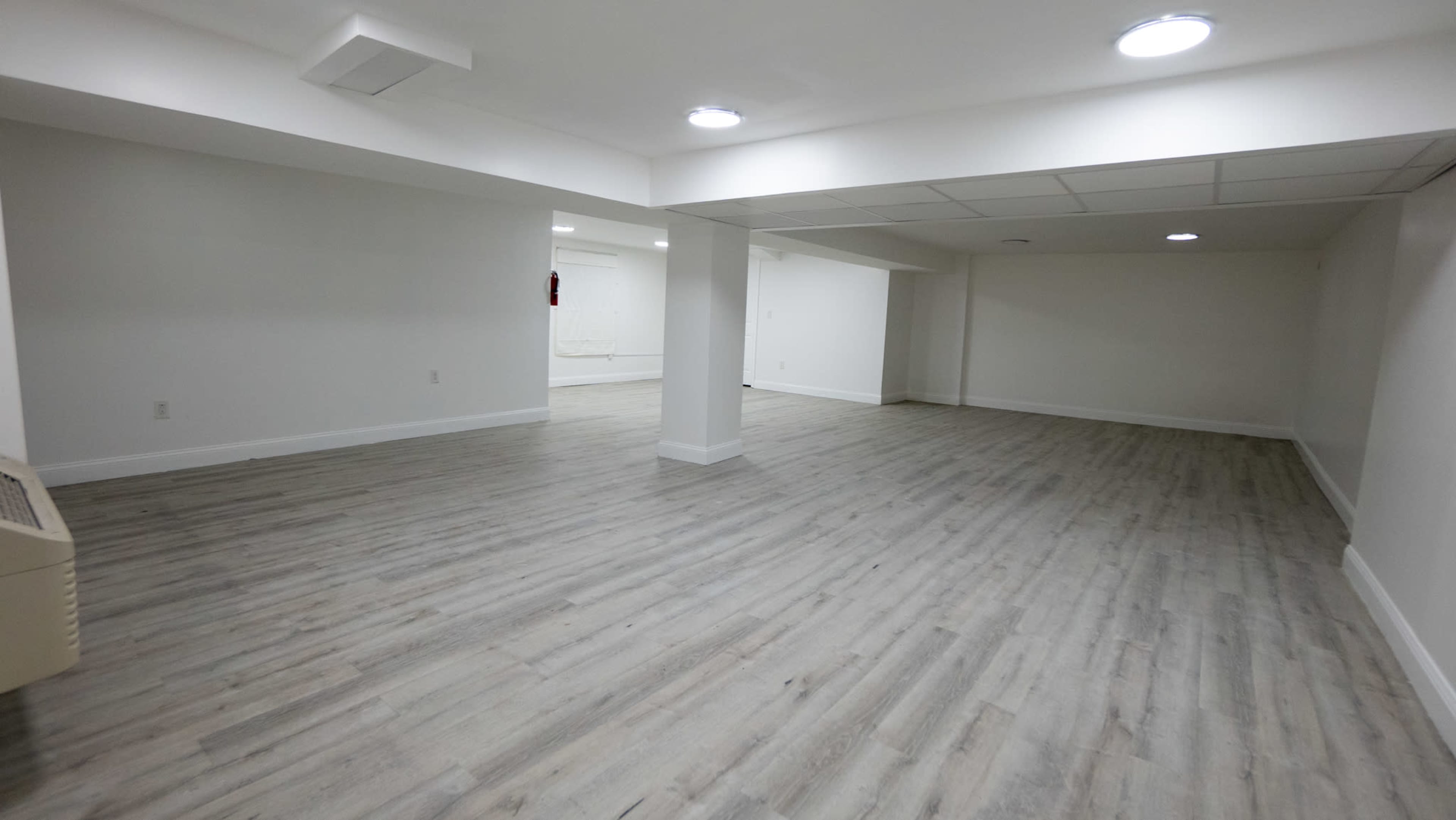 A large empty room with light-colored flooring and white walls, illuminated by ceiling lights.