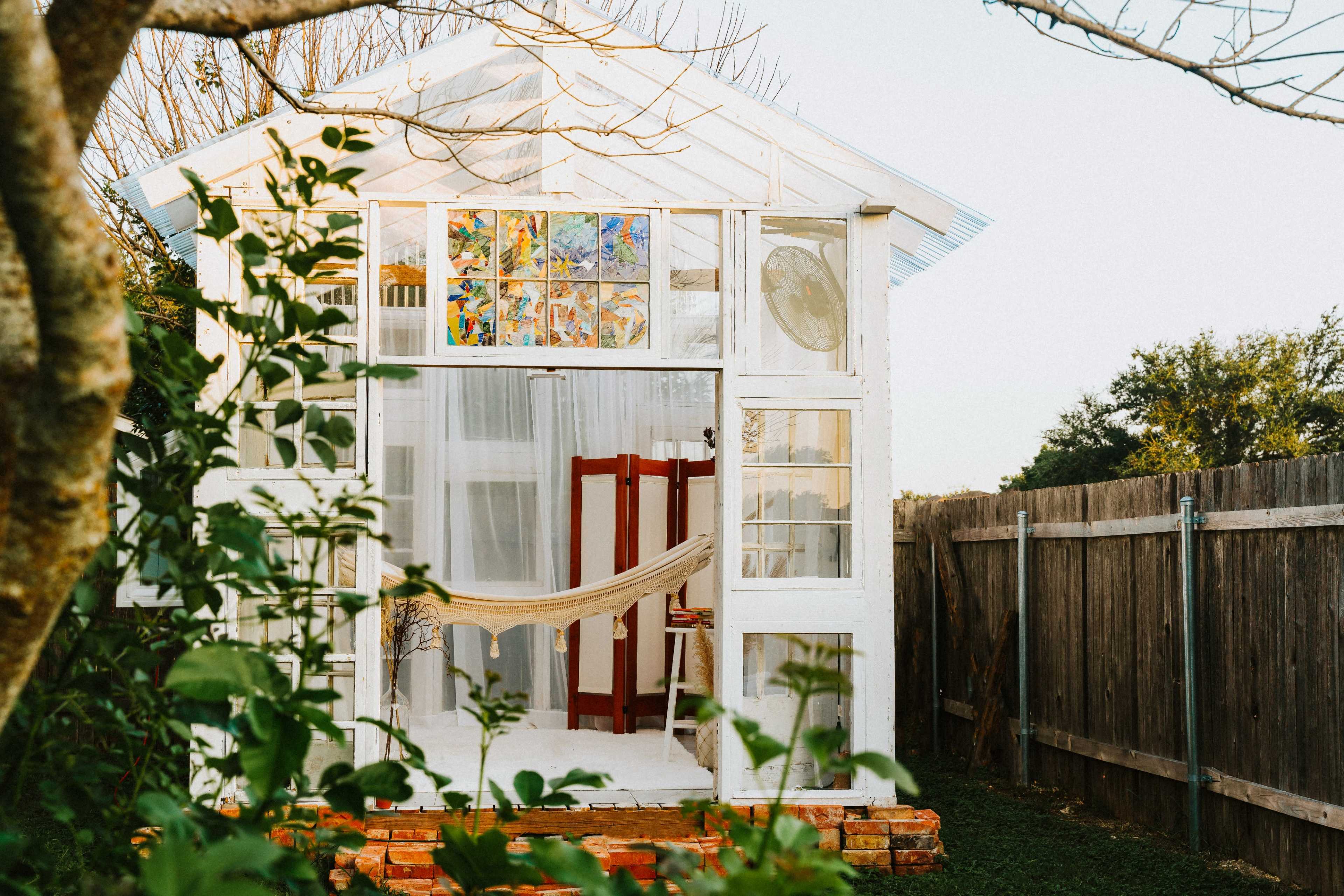 A small white shed with colorful stained glass windows is surrounded by greenery and features a hammock hanging in the entrance.