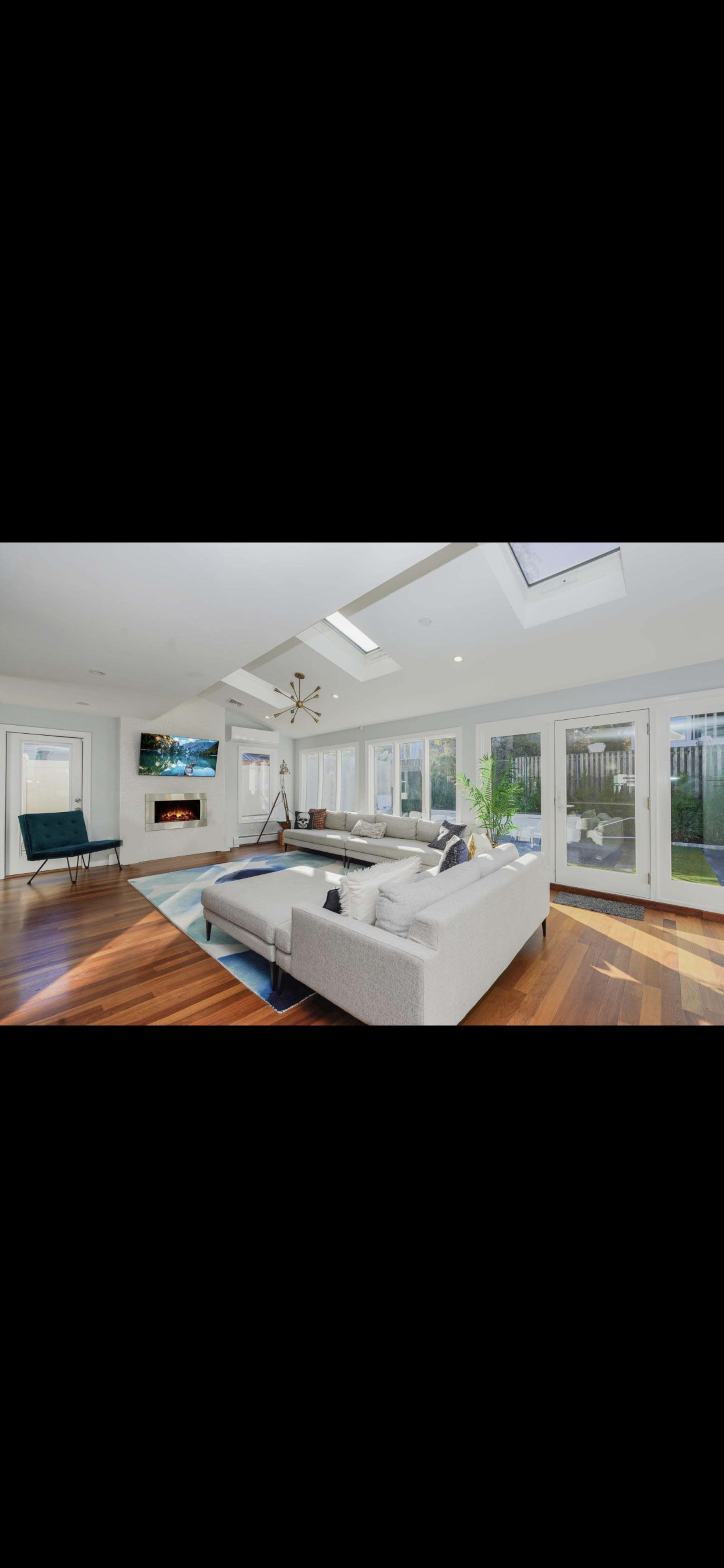 The image depicts a modern living room with a large white sectional sofa, a flat-screen TV mounted on the wall, and large windows providing natural light.