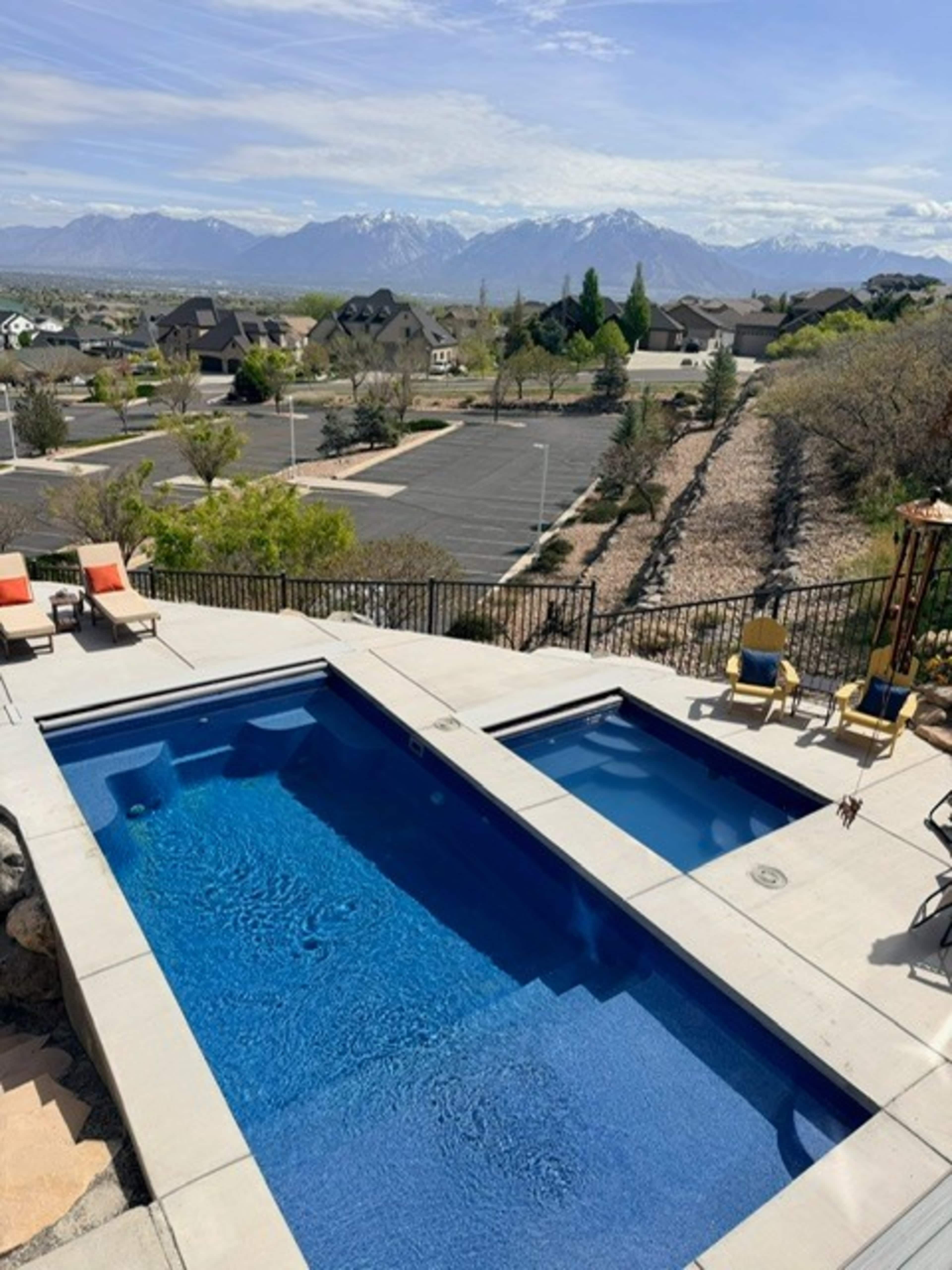 The image shows a modern swimming pool area overlooking a neighborhood and distant mountains under a clear blue sky.
