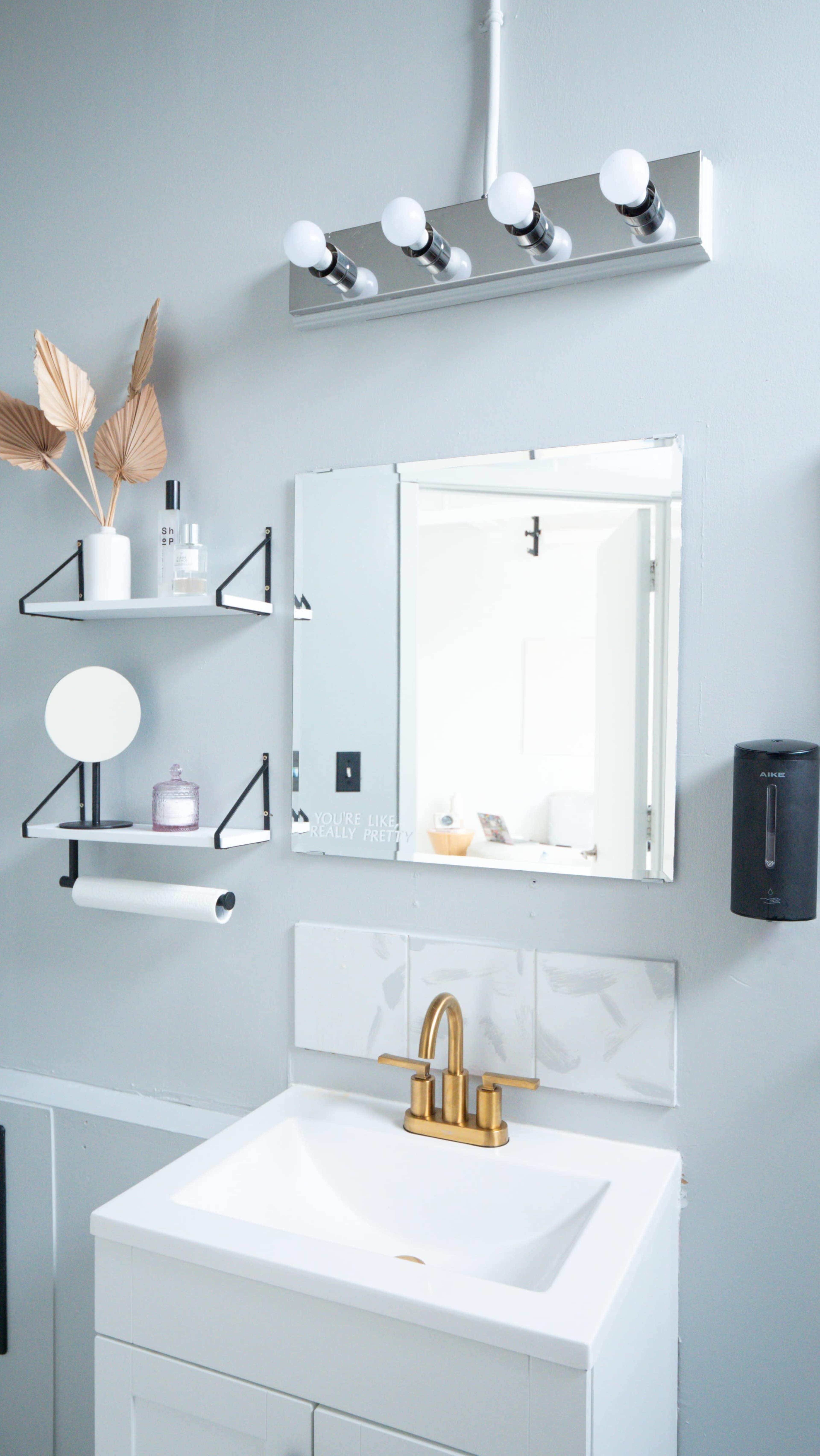 A modern bathroom with a mirror, a light fixture with four bulbs, a sink with a gold faucet, and decorative elements like a plant and skincare products.