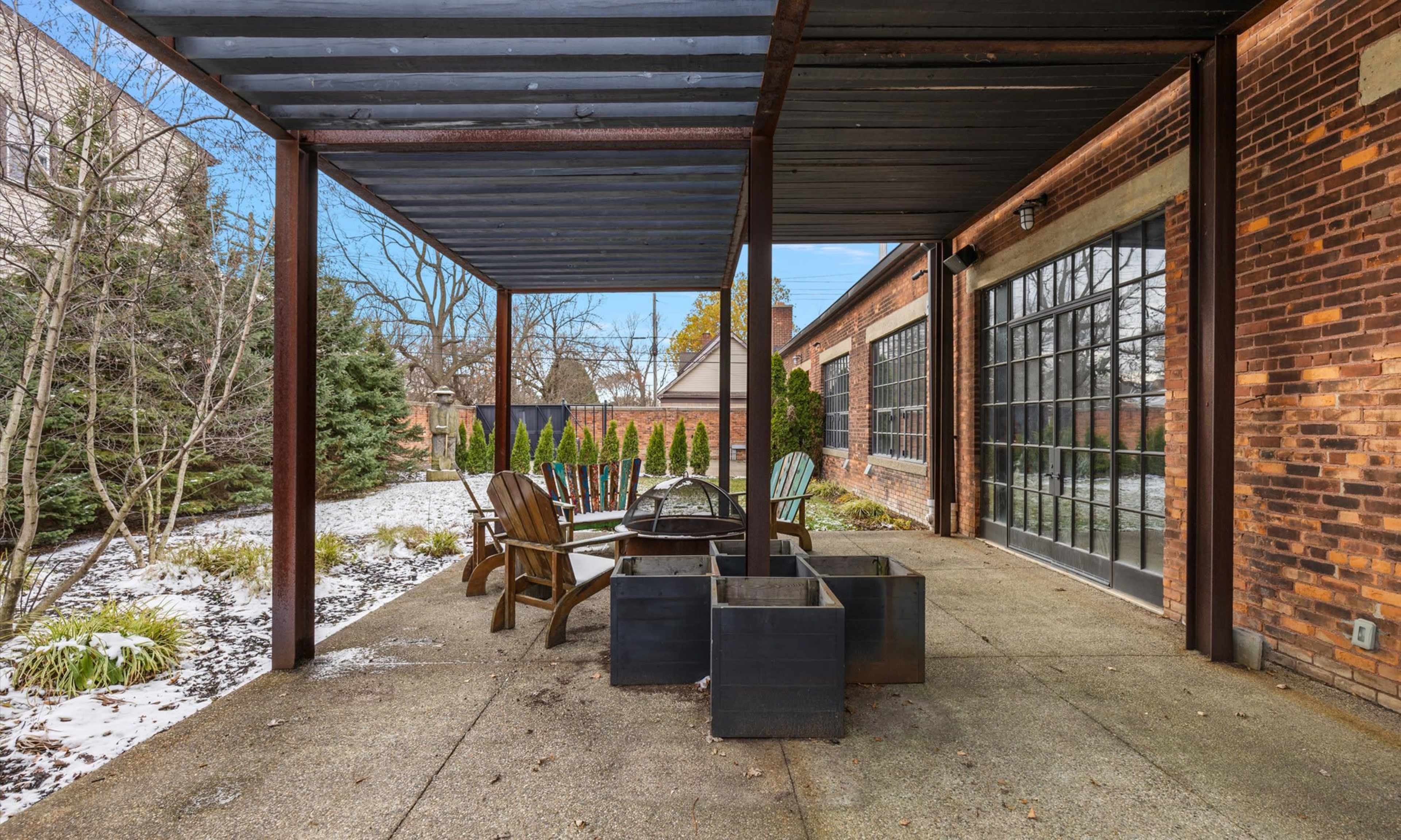 The image depicts a patio area with a covered seating arrangement, including wooden chairs and metal planters, surrounded by a landscaped yard.