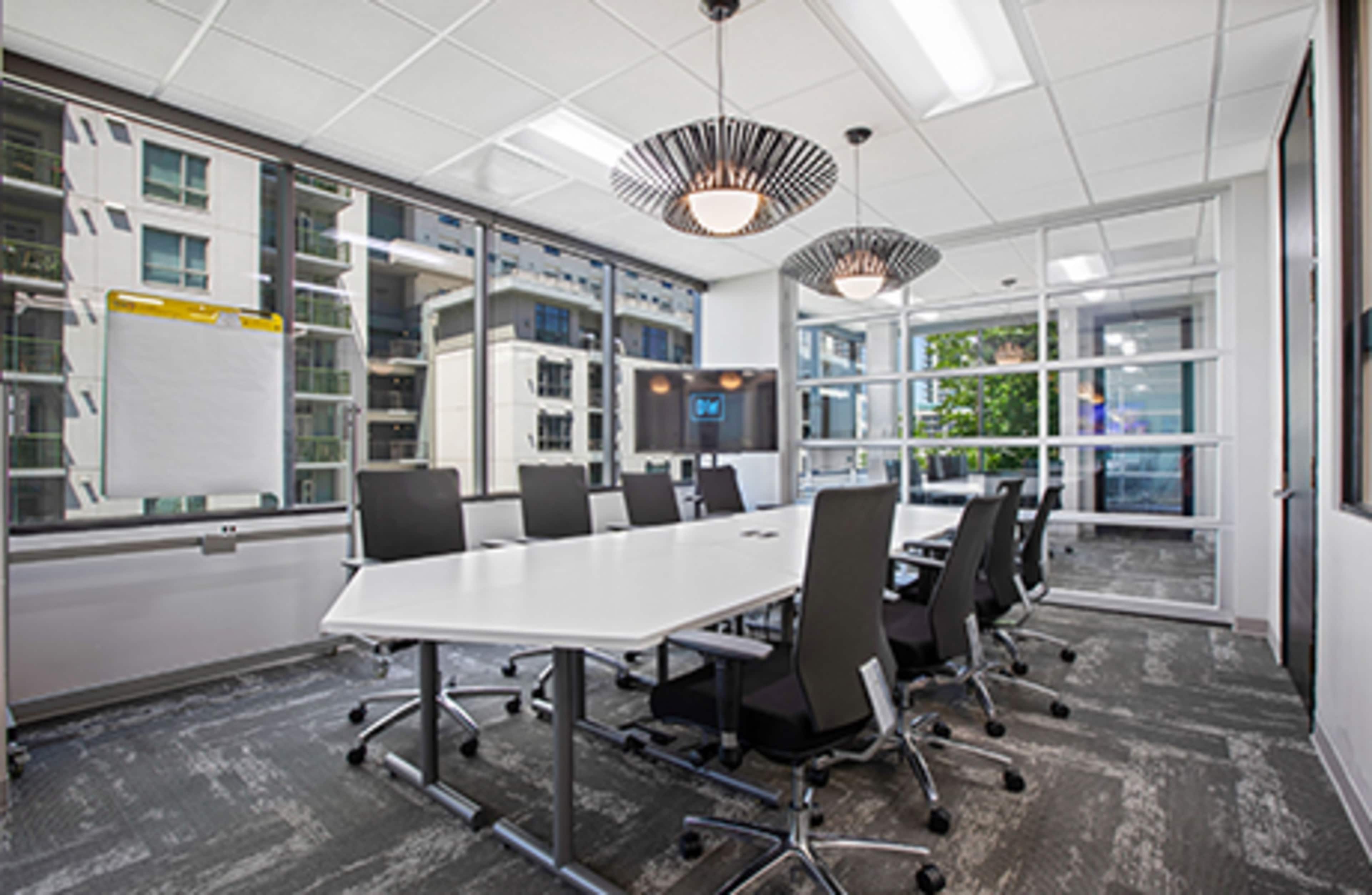 A modern conference room with a large white table, black chairs, and floor-to-ceiling windows overlooking an urban landscape.