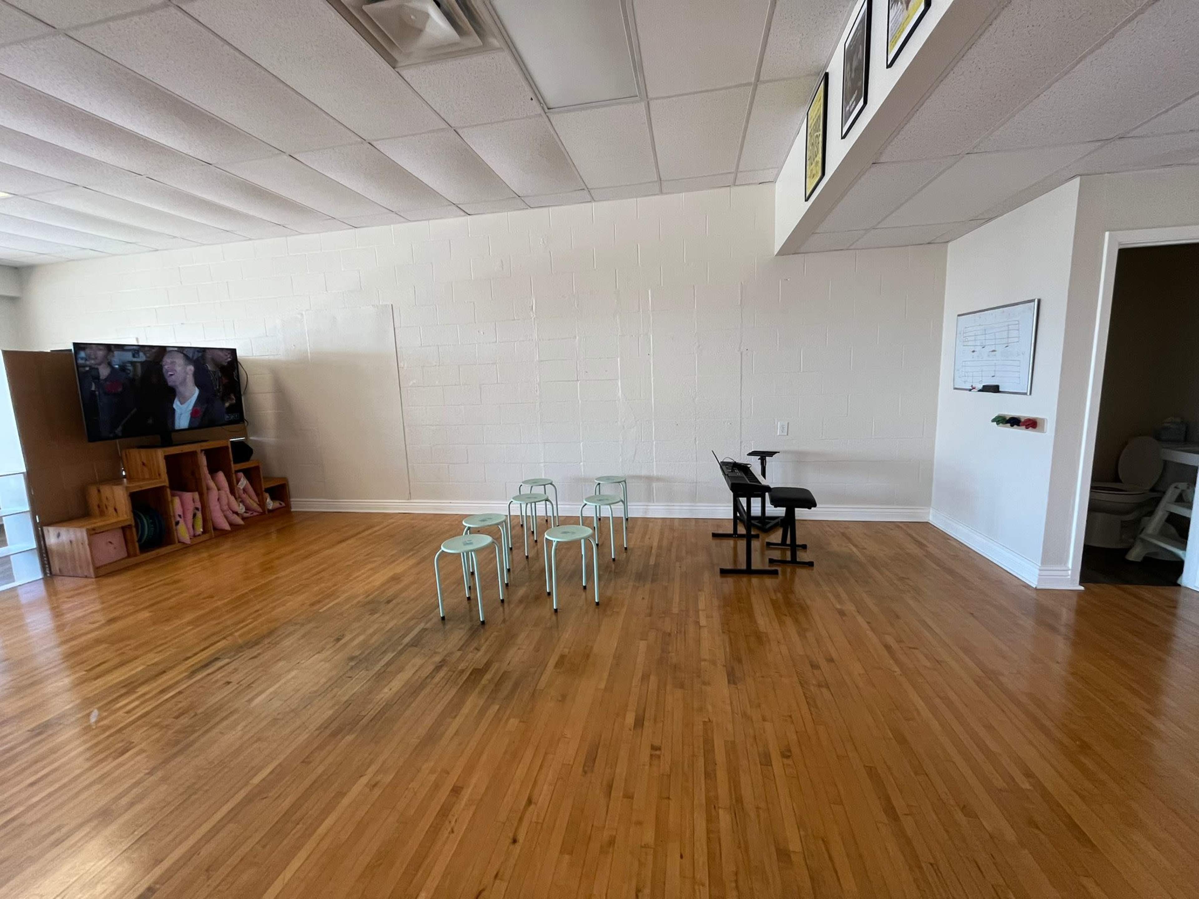 The image shows a spacious, bare room with wooden floors, featuring a small table and several chairs arranged in the center, a television on one wall, and a doorway leading to another room.