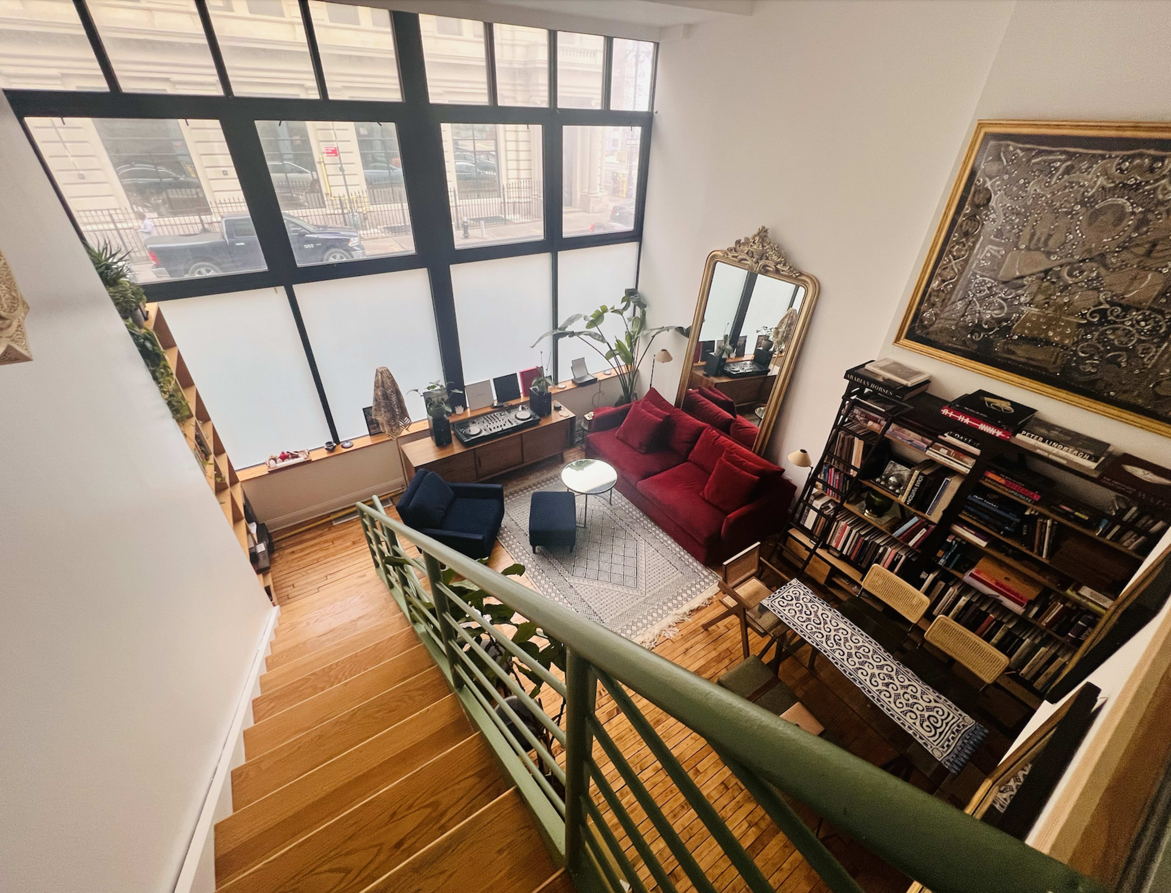 A staircase leads down to a cozy living area with a red sofa, a coffee table, and a bookshelf, all illuminated by large windows.