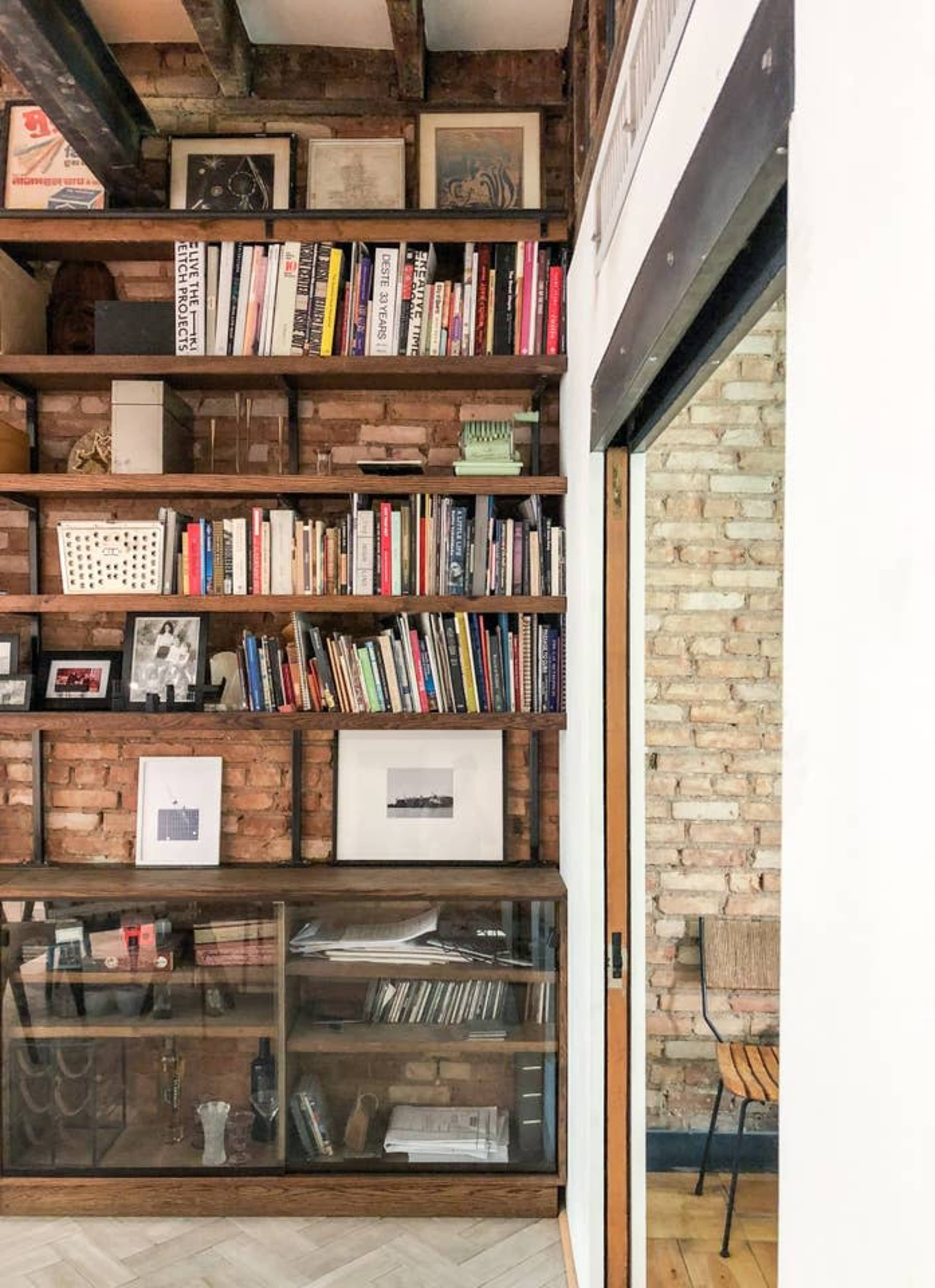 A wooden bookshelf filled with books and framed photographs in a room with exposed brick walls.