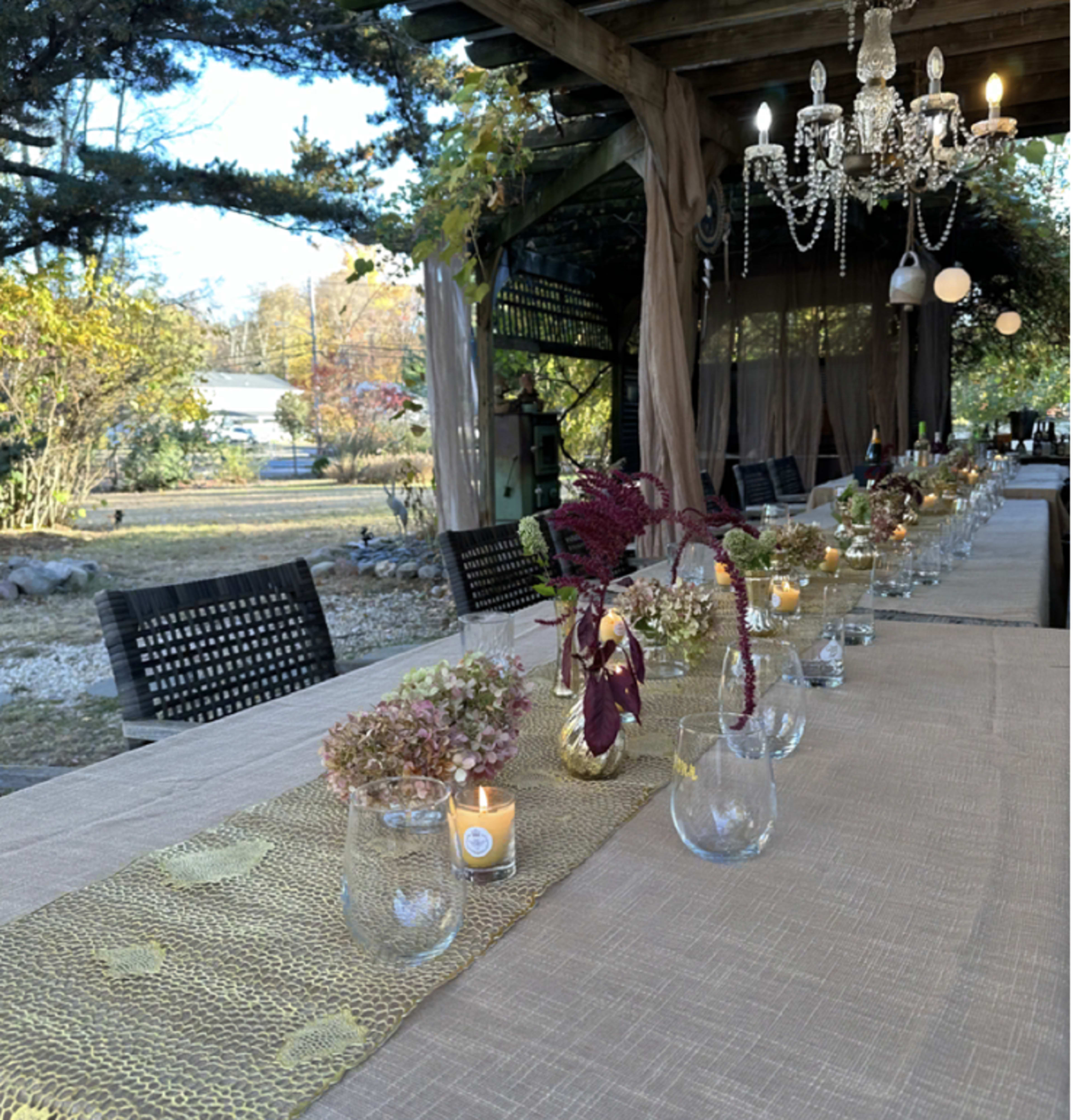 A long outdoor table is set with glassware, candles, and floral arrangements under a wooden structure adorned with a chandelier.