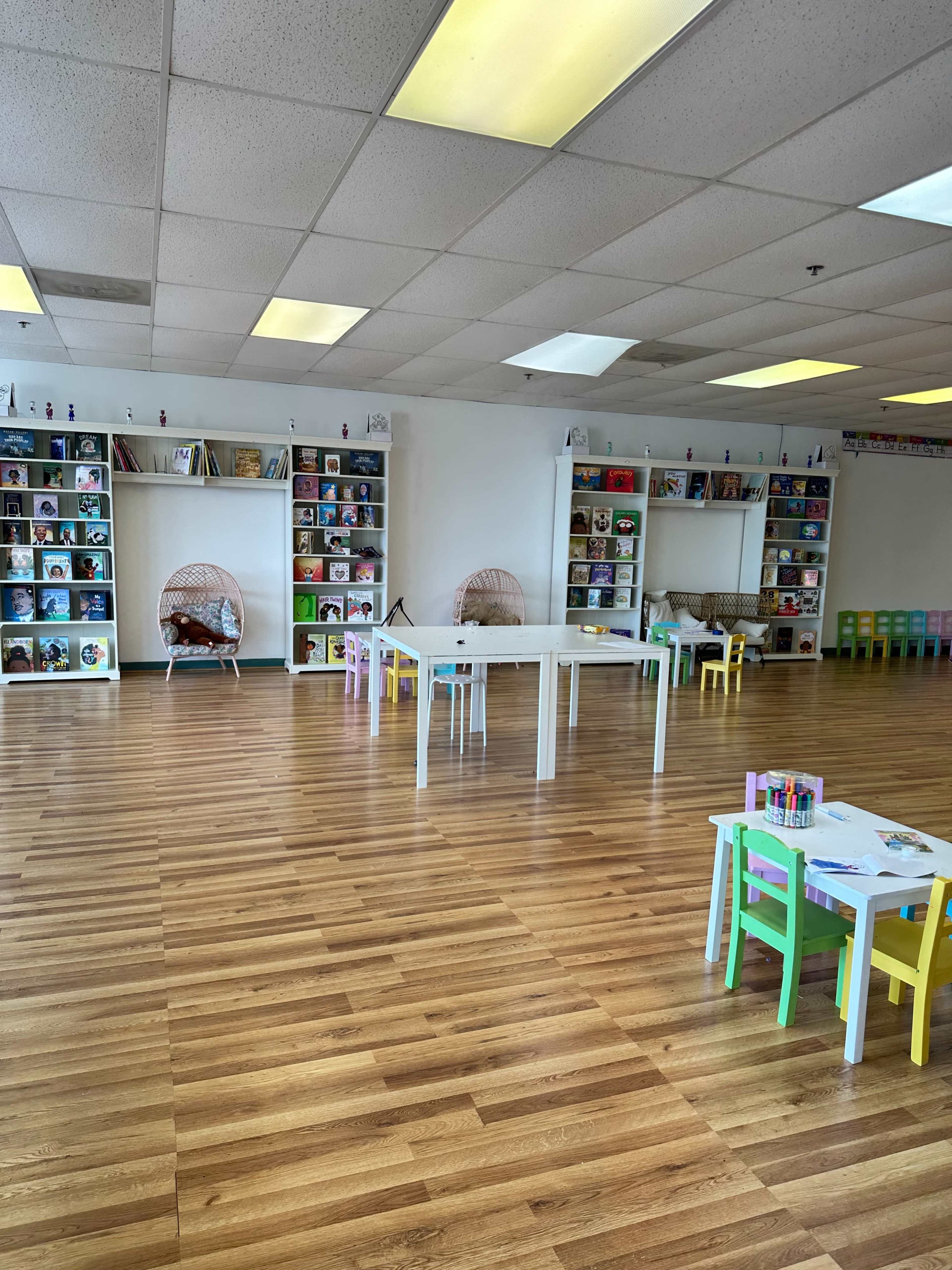 The image shows a spacious children's reading area with white shelves filled with books and several small tables and chairs arranged on a wooden floor.