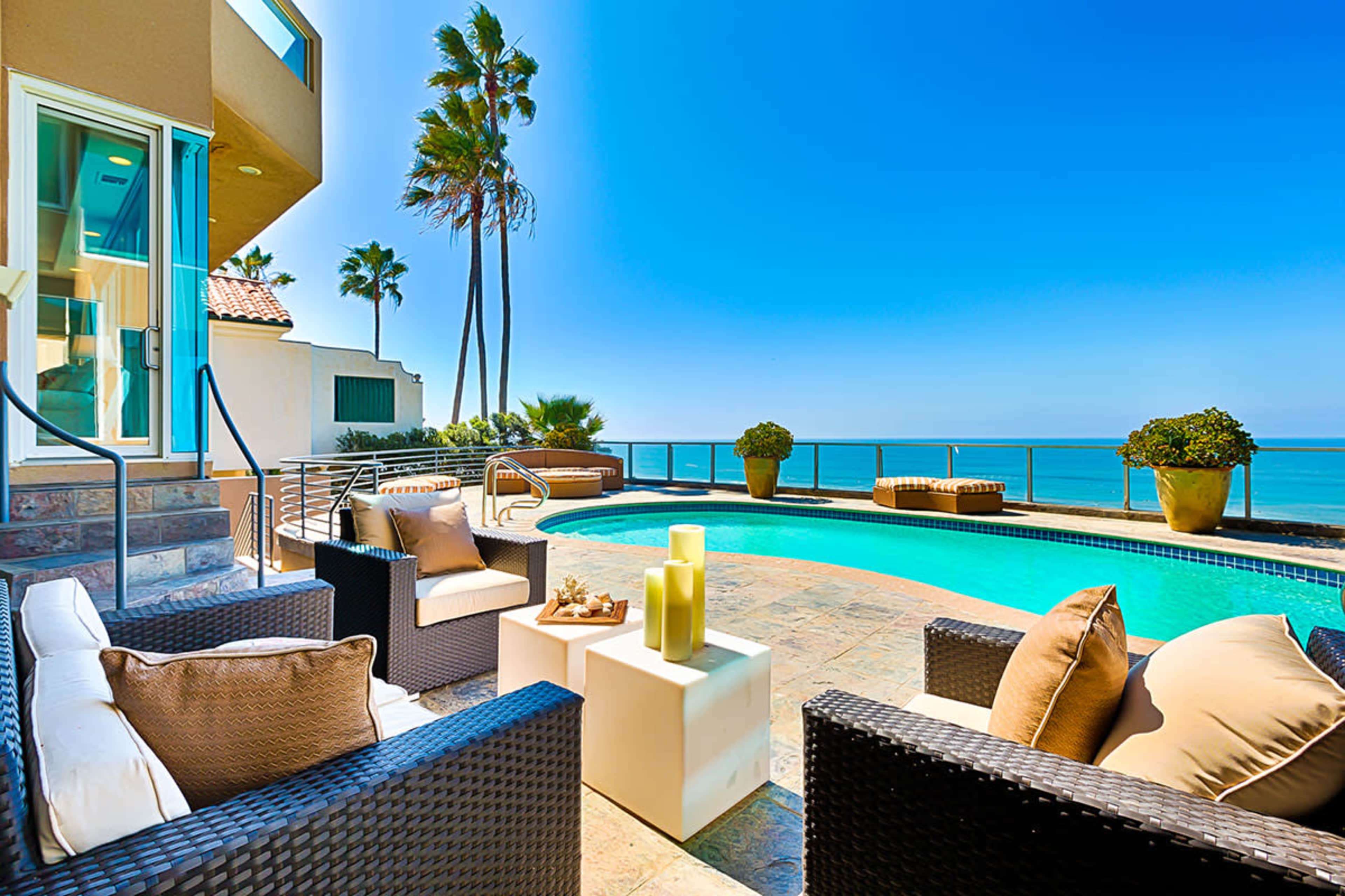 A patio area featuring wicker seating and a small table overlooks a swimming pool beside the ocean, framed by palm trees under a clear blue sky.