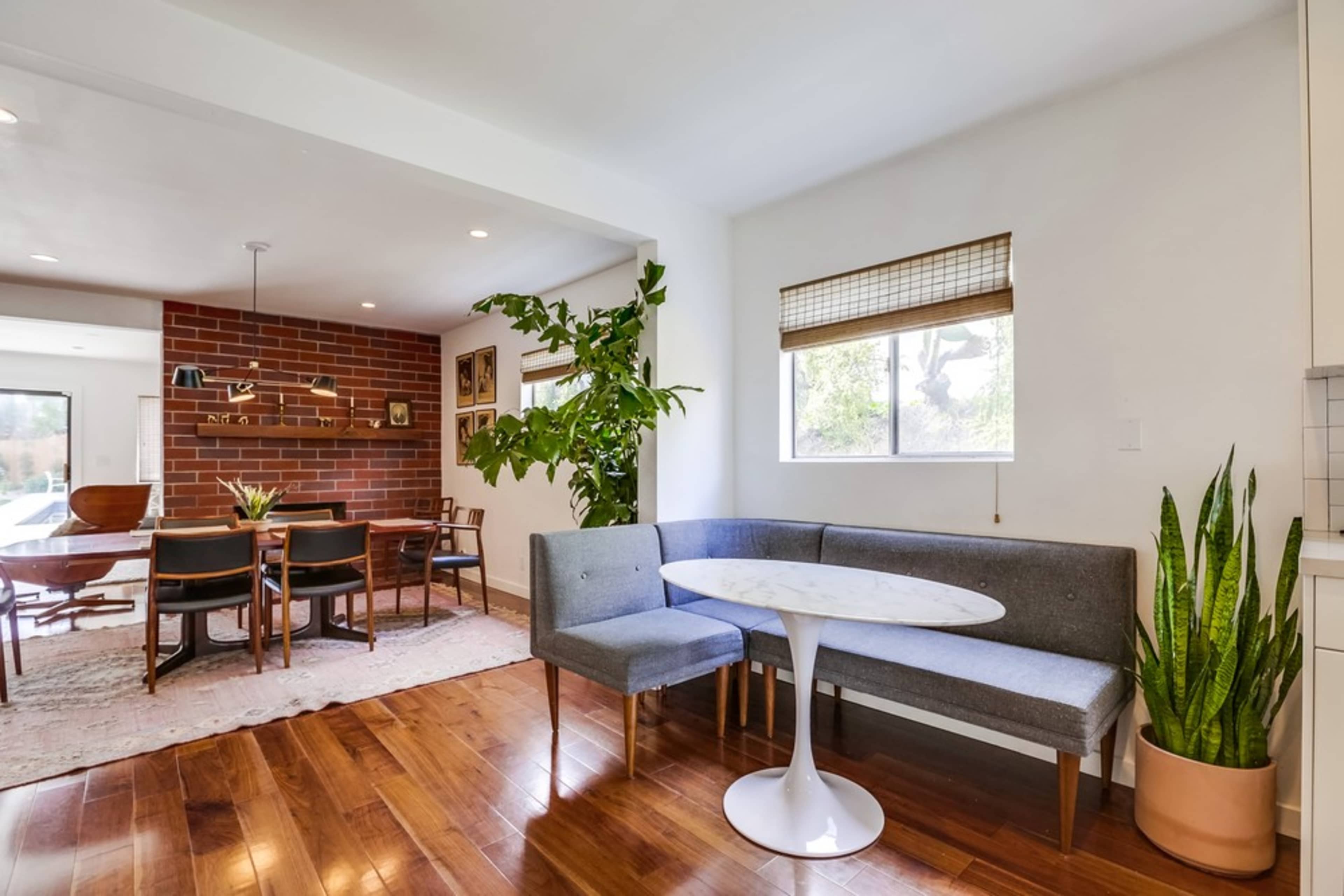 A modern dining area features a round table with a white top and a gray upholstered bench surrounded by houseplants and hardwood floors.