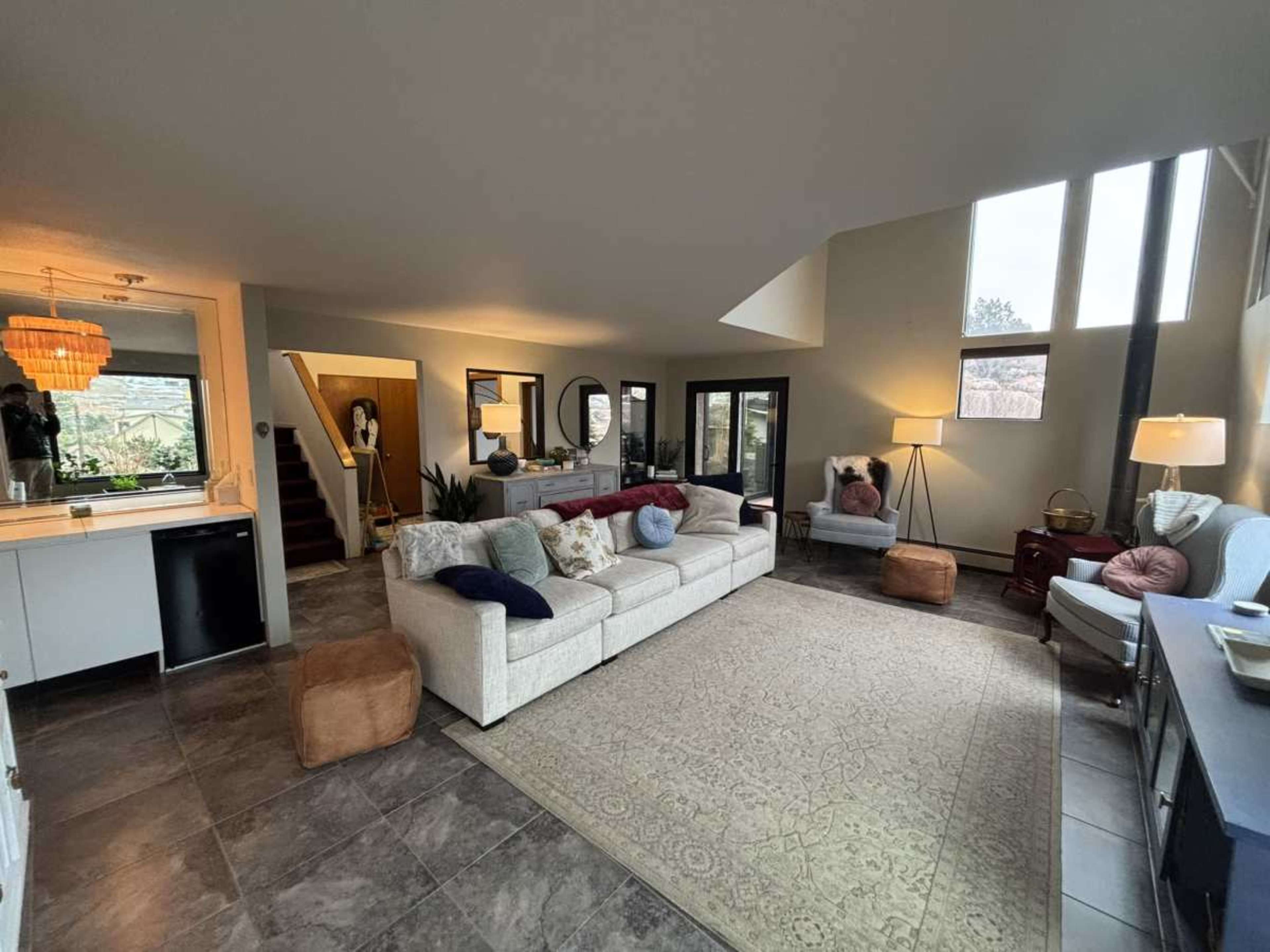 The image shows a spacious living room with a large sectional sofa, a patterned area rug, large windows, and a view of a staircase leading to another level.