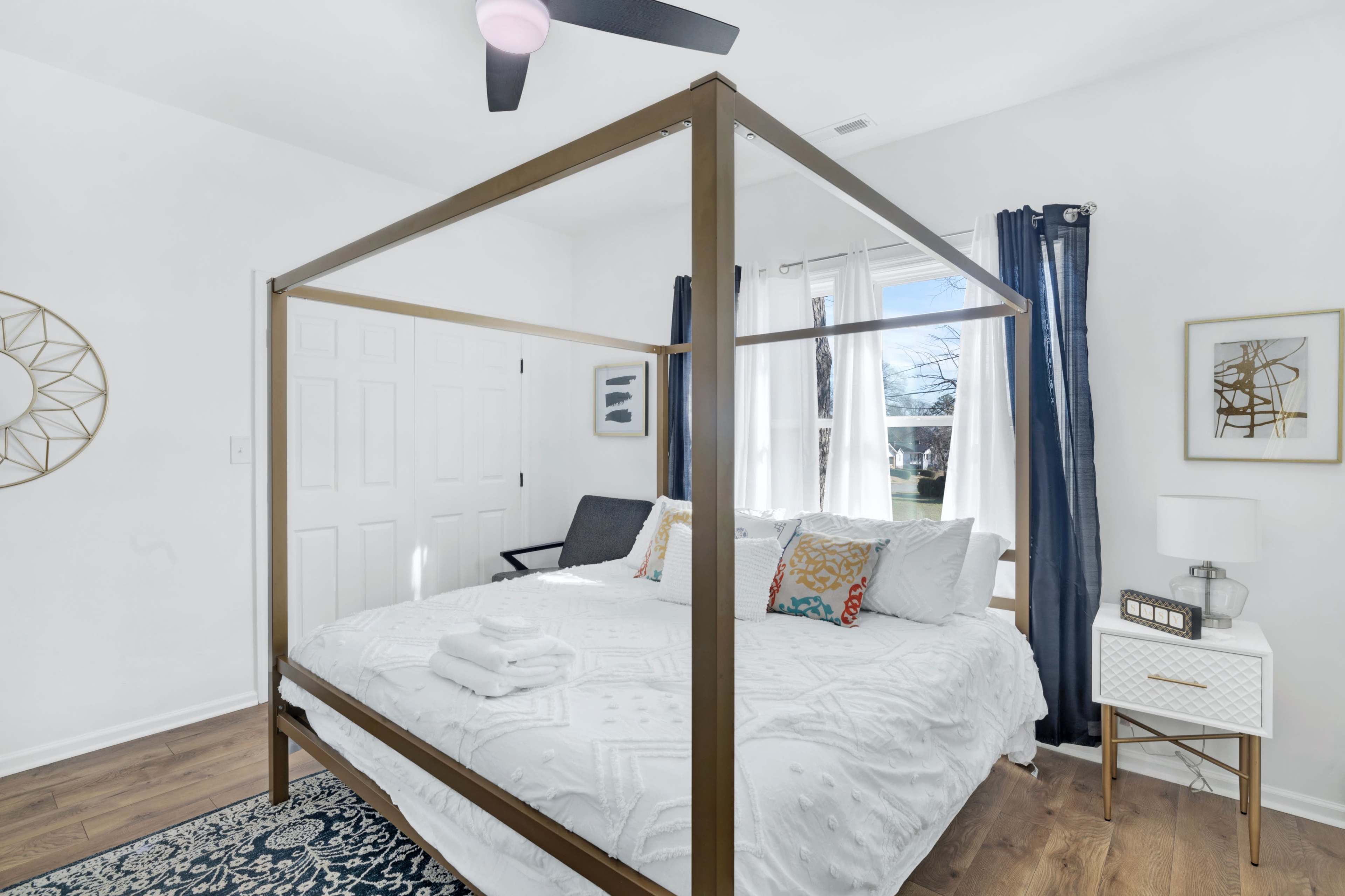 A modern bedroom features a four-poster bed with white bedding, a nightstand, and a window with blue curtains.