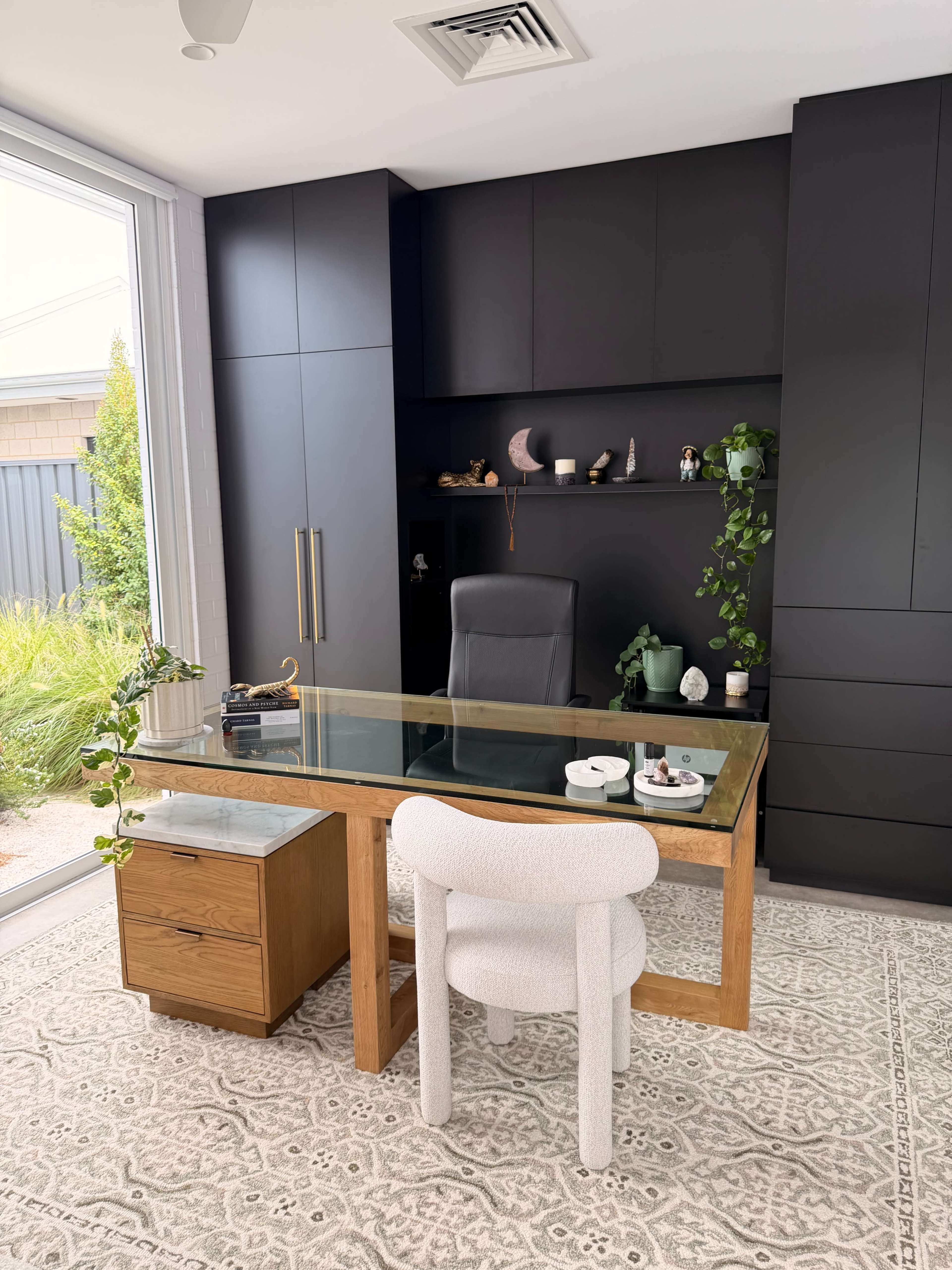 The image shows a modern home office featuring a glass-top desk with a wooden drawer unit, a black office chair, and shelves displaying decorative items against a backdrop of greenery outside.